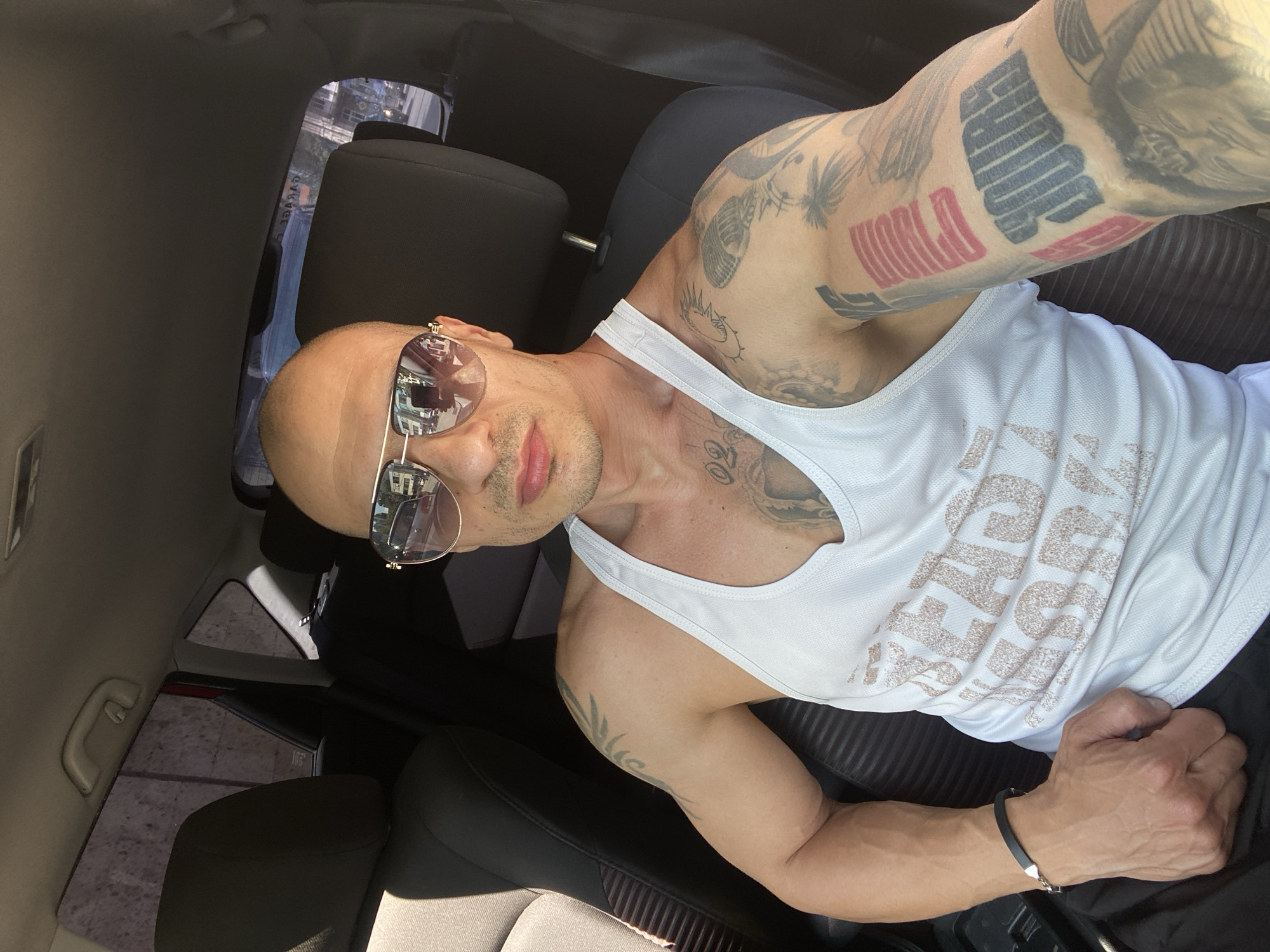 The image displays a man taking a selfie inside a car, with the photo oriented 90 degrees clockwise. The subject is a bald man with light stubble, wearing reflective aviator sunglasses and a white tank top. He presents a neutral, confident expression.
His arms are heavily tattooed. On his left arm (viewer's right), large block letters are visible, with "WORLD" clearly in red, and black text above it that appears to be "FAMOUS," set among other black graphic tattoos. His right arm (viewer's left) also features various black tattoos, including abstract designs and a sun-like symbol. The white tank top has light brown or gold text across the chest, partially visible, possibly reading "EAGLE." A black bracelet with a silver clasp is worn on his left wrist.
The setting is a car interior, characterized by black seats and light grey/beige ceiling and door panels. Bright daylight, evident from reflections in the sunglasses and the lighting through the windows, suggests a sunny daytime. Reflections in his sunglasses show urban buildings. Through the rear window, a reversed sign reading "GARAGE" is faintly visible, along with a street or parking lot and other structures, indicating an urban or suburban environment. The scene depicts a casual moment within a vehicle, likely in Pleven, Bulgaria.
