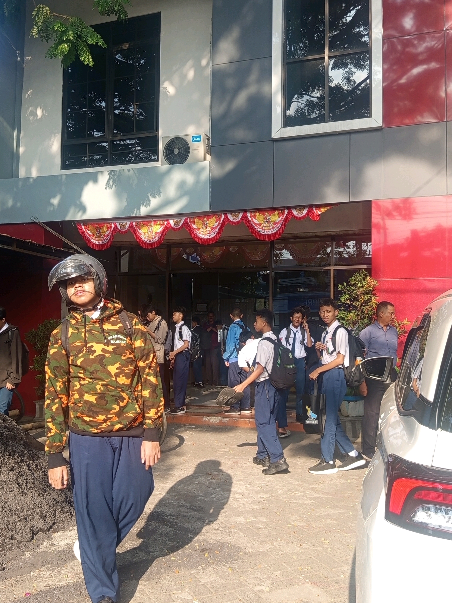 The image depicts a group of students, many in school uniforms, gathered outside a building. The building features a grey and red facade with large windows reflecting trees. A red and white decorative banner with a national emblem is visible above the entrance. The foreground is dominated by a pile of dark aggregate material and a white car partially visible on the right.
A young person wearing a camouflage hoodie, dark blue pants, and a clear helmet is prominently positioned in the lower left of the frame, facing the camera and casting a shadow. Other students, mostly wearing white shirts and dark pants or shorts, are seen in various groupings, some with backpacks. One student is making a hand gesture. The overall impression is of a school environment, possibly during a break or at the end of the school day. The presence of an air conditioning unit on the building suggests an urban or suburban setting. While the image does not explicitly state the location, the banner with the Garuda Pancasila emblem indicates it is within Indonesia. Given the context of schools and the visible architectural style, it is plausible this scene is located in the city of Malang, Indonesia.