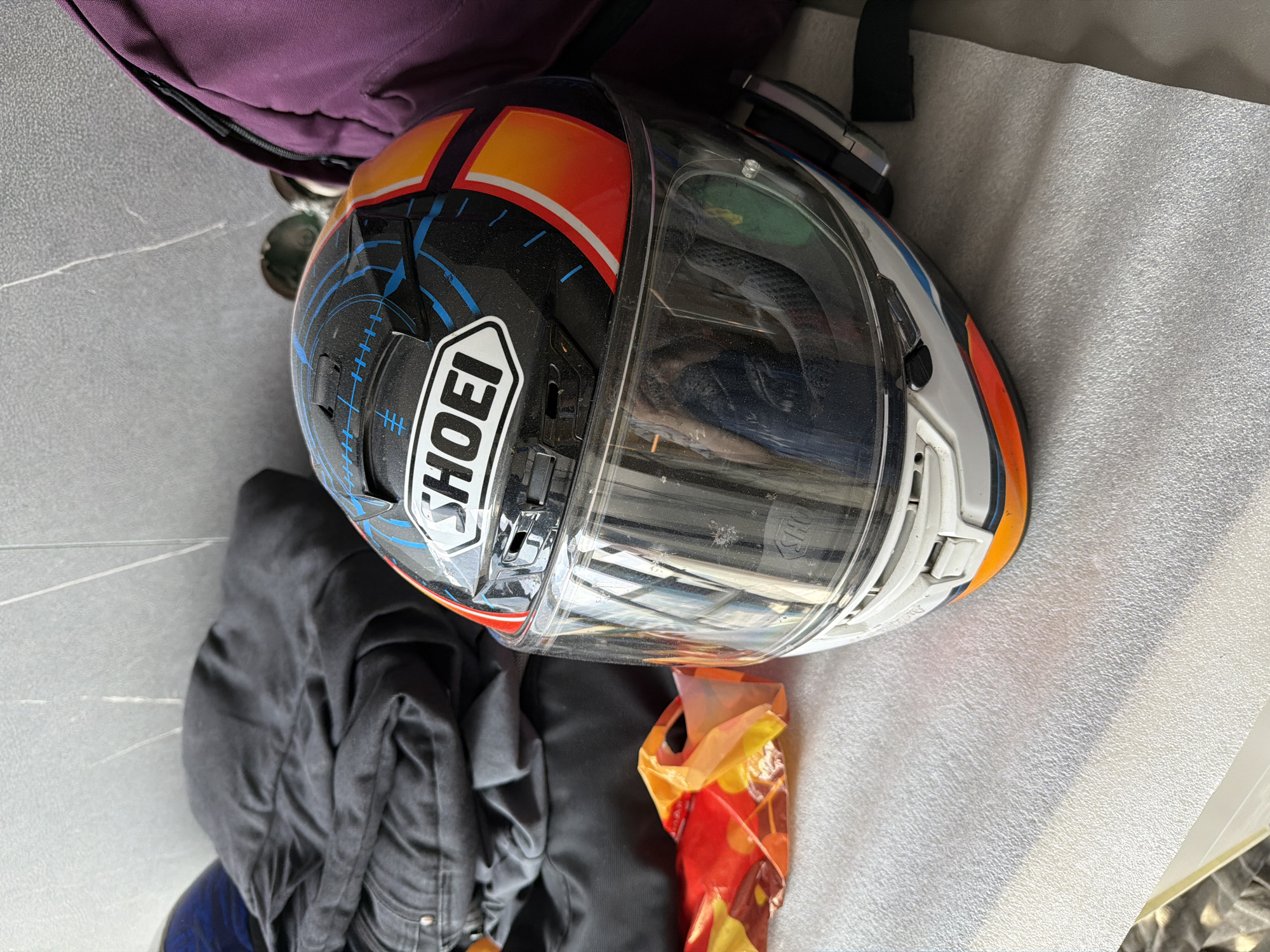The image depicts a Shoei motorcycle helmet with a clear visor, resting on a textured surface. The helmet is predominantly black with orange and white accents, and a Shoei logo is visible on the side. Adjacent to the helmet is a crumpled bag with a red and yellow pattern, a partially visible black jacket, and a glimpse of a purple bag. The setting appears to be indoors. There is no evidence in the image to confirm that the location is Khimki, Russia.