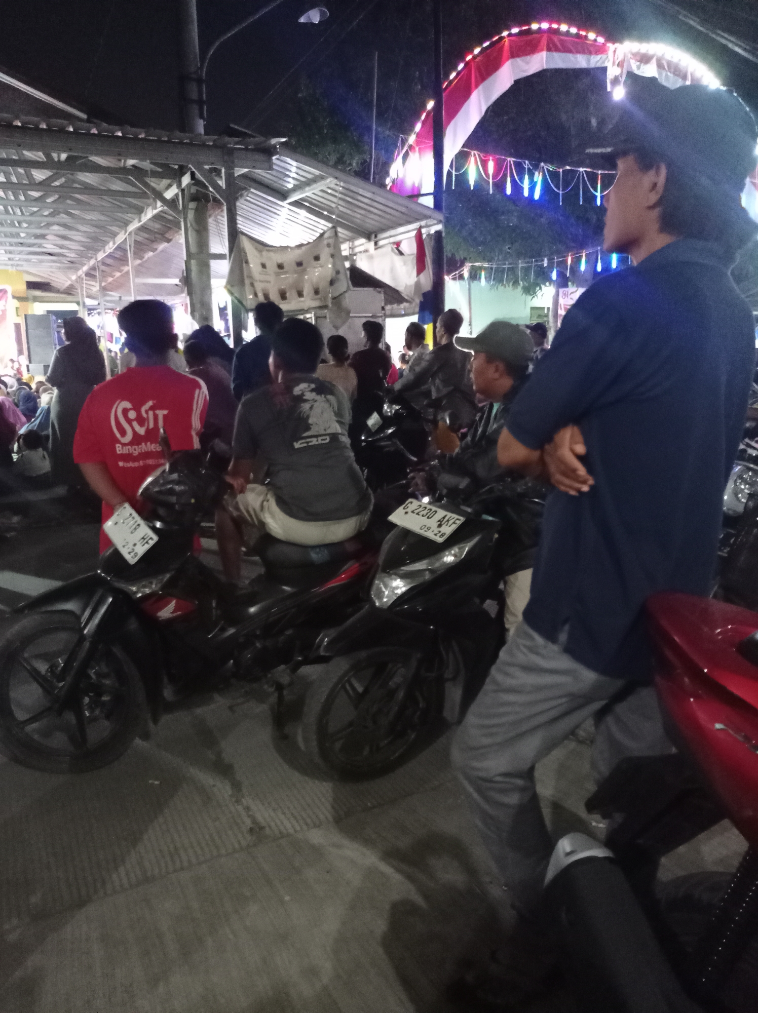The image depicts a nighttime scene in Plumbungan, Indonesia, likely at a local event or gathering. In the foreground, several motorcycles are parked, with their license plates visible. One motorcycle's plate reads "G 2718 HF" and another "C 2230 AKF". A man wearing a dark blue t-shirt and grey pants stands with his arms crossed, facing away from the camera. Behind him, another man in a grey t-shirt and light-colored shorts is seated on a motorcycle. To the left, a person in a red t-shirt with a white logo that says "BanggaMuda" is visible. Other individuals and motorcycles are present in the background. The scene is illuminated by overhead lights, festive colored lights strung across a structure, and the glow from a red motorcycle fender in the lower right corner. The overall atmosphere suggests a community event or market taking place after dark.