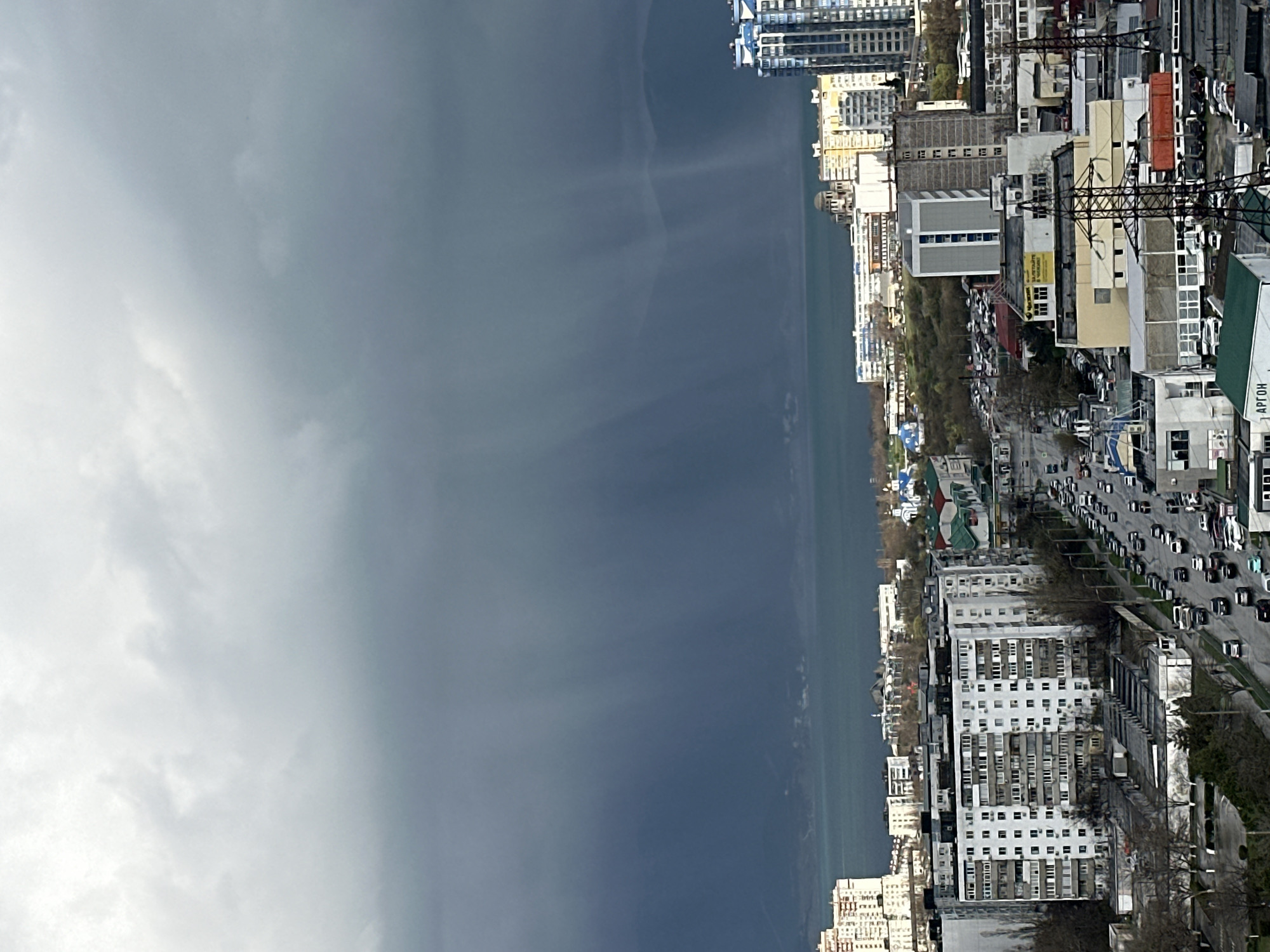 The image shows an urban environment with a mix of natural and man-made elements. A cloudy sky covers a significant portion of the frame. The line of the ocean and the city is partially obscured by the clouds. The buildings are of various heights and architectural styles. A road with moving vehicles runs along the right side of the image, near the city. The overall impression is a dense, bustling city.