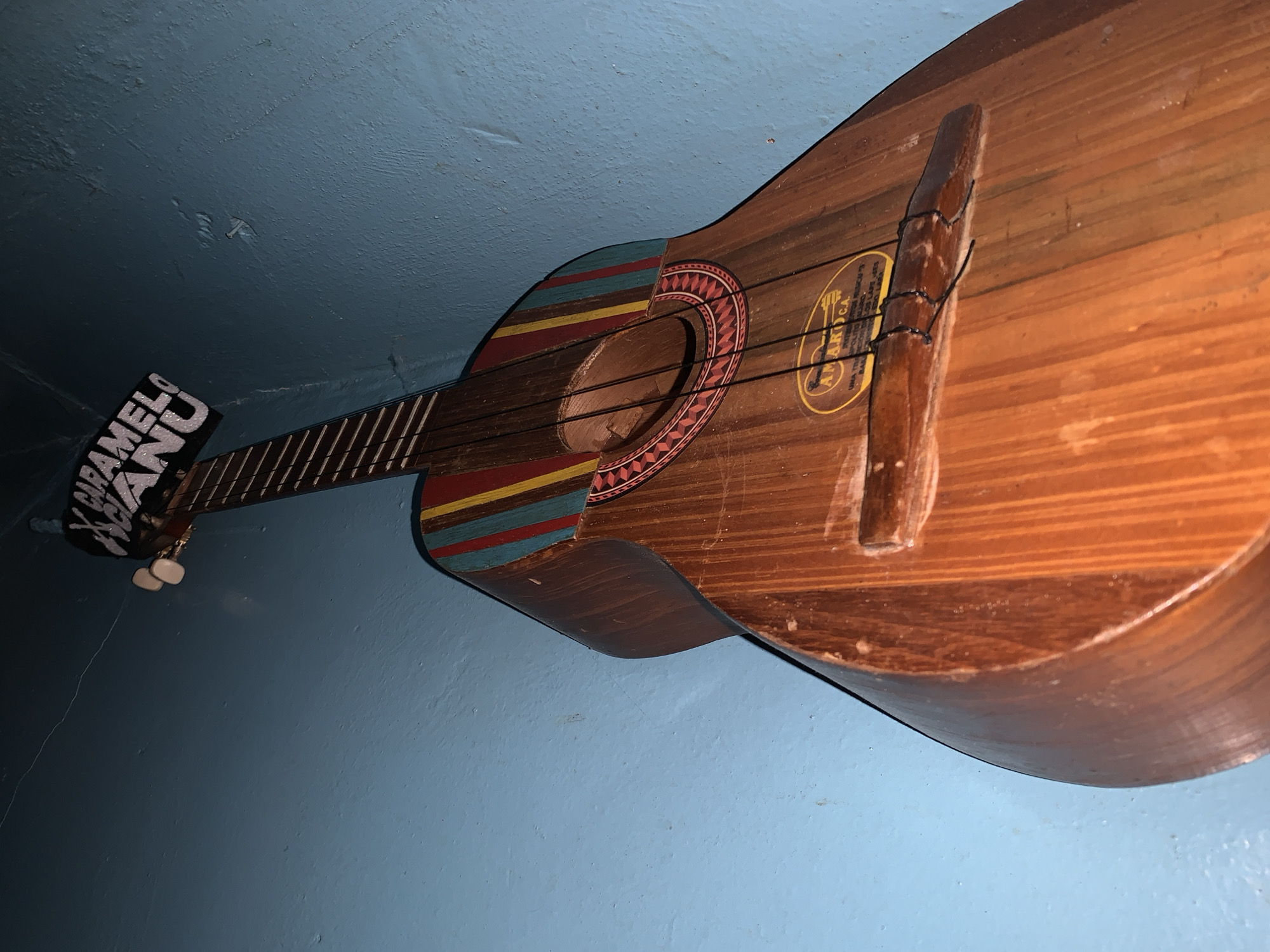 A stringed musical instrument, resembling a small guitar or a Venezuelan cuatro, hangs vertically against a textured light blue background, likely a wall or ceiling. The instrument, crafted from wood with visible grain, is adorned with vibrant, traditional-style patterns. Around the sound hole, a circular design in red, black, and white is prominent. The upper bout features horizontal stripes in red, yellow, blue, and black.
The instrument has a wooden bridge, to which black strings are tied, though some appear missing or loose. A yellow oval label is affixed to the instrument's body, above the bridge. The headstock, visible at the top-left, has two white tuning pegs.
**Visible text:**
On the headstock, the phrase "CARAMELO MANU" is boldly displayed in white capital letters against a dark background.
The yellow oval label on the instrument's body clearly shows "AMAROCA" at the top. Below it, smaller text is visible, including "UNA TRADICION DE LUJOS C.A." and "BARQUISIMETO", which is a city in Venezuela.
The background is a consistent light blue with an uneven, bumpy texture, possibly stucco or poorly painted concrete. The lighting is somewhat dim, creating shadows that emphasize the texture of both the instrument and the background. The instrument appears to be an older, rustic piece, possibly handmade, displayed as an artifact or decoration. The overall impression is of a culturally significant object from Santa Ana, Venezuela.