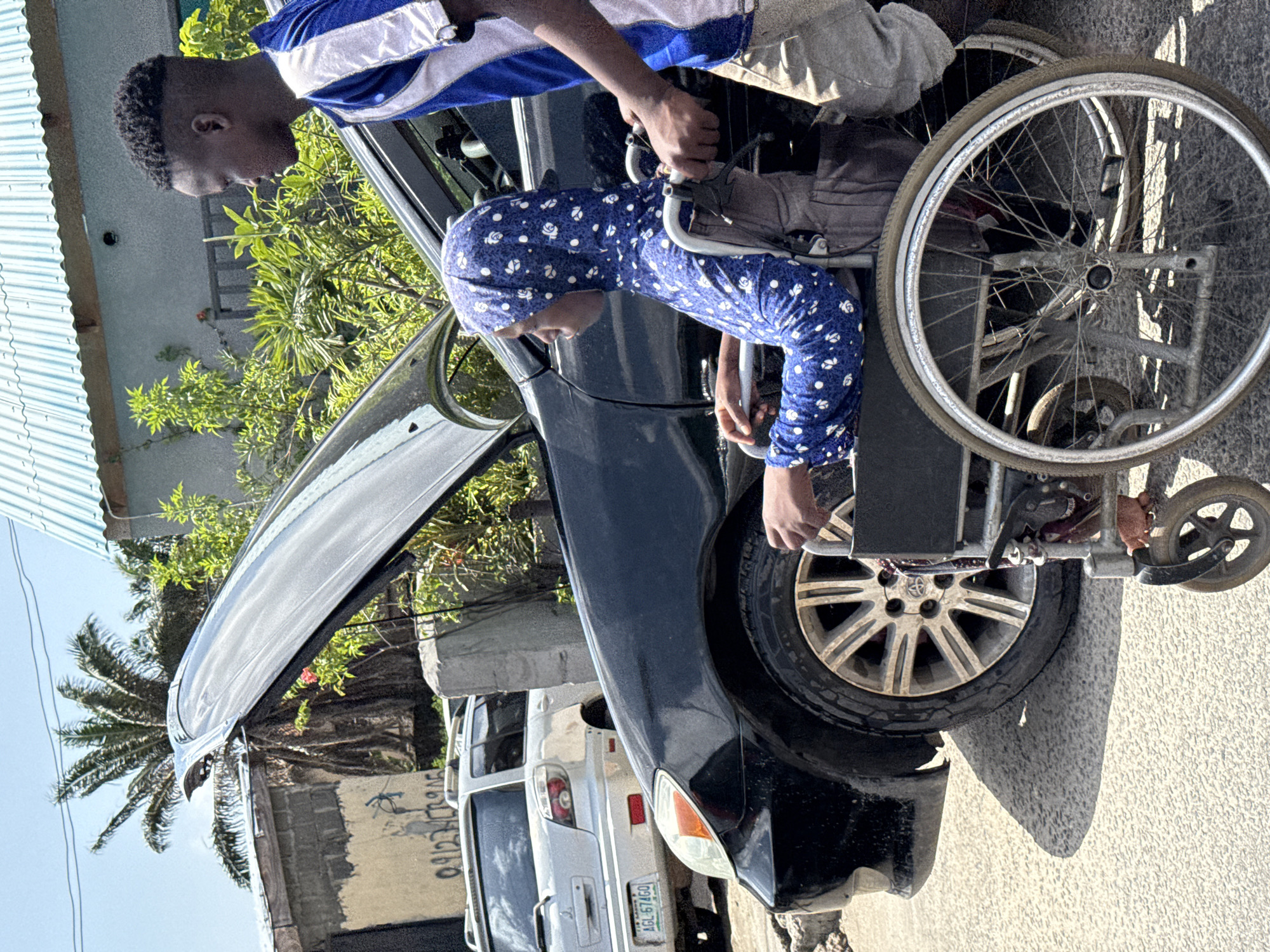 The scene is set on a street in Ado, Nigeria. A black car is parked on the roadside with its hood open. A young person in a blue and white patterned long-sleeved shirt and head covering is seated in a wheelchair next to the car's open door. An individual wearing a blue and white athletic shirt is assisting the person in the wheelchair, possibly helping them transfer from the wheelchair into the vehicle. A white car is parked behind the black car. Vegetation, including a palm tree, and parts of buildings are visible in the background. The asphalt street is visible in the foreground. A vehicle license plate with the letters AGL and numbers 67460 can be seen on the white car.