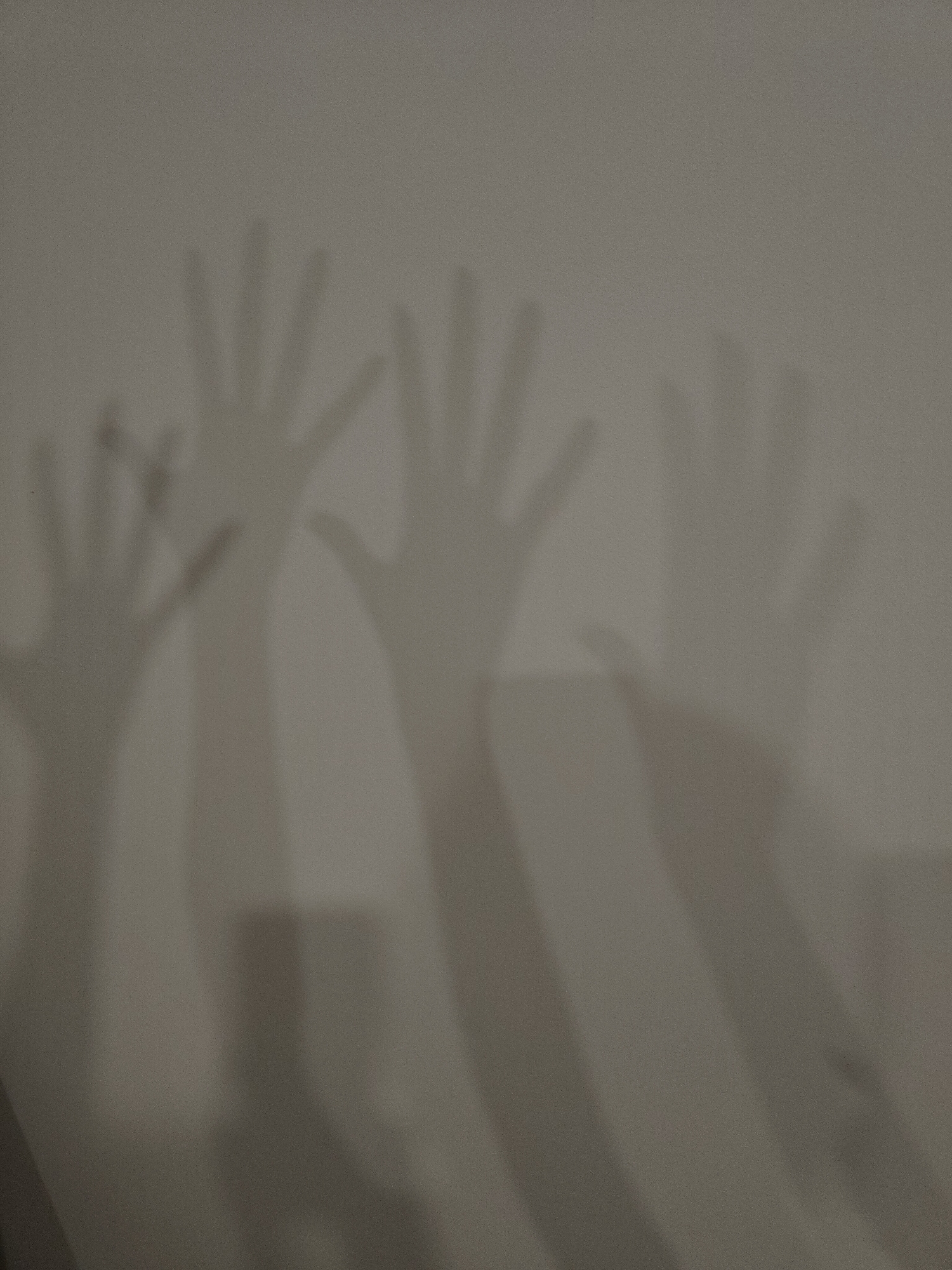The image features the shadows of multiple hands and arms cast upon a light-gray textured wall. At least four distinct hand shadows are visible, predominantly in the upper and middle sections of the frame. One smaller hand shadow is present on the far left, with its fingers spread upwards, next to a larger, more defined hand shadow. Towards the center and right, additional hand shadows, varying in clarity and size, are visible, some appearing softer or partially obscured. The arm shadows extend downwards, sometimes overlapping, and lead off the bottom edge of the frame. The hands generally show fingers extended or slightly spread, suggesting gestures such as waving, reaching, or simply being held up.
The activity depicted is the creation of these shadows, implying one or more individuals are positioned between a light source and the wall. The strong illumination creates distinct, elongated forms. The setting appears to be indoors, likely in a room with a plain wall, as no specific furniture or outdoor elements are visible. The time of day is not explicitly indicated, but the presence of clear shadows suggests an artificial light source or a specific angle of natural light. The overall color palette is monochromatic, dominated by shades of gray and light brown, which lends an abstract and somewhat mysterious atmosphere to the scene. There is no visible text in the image.