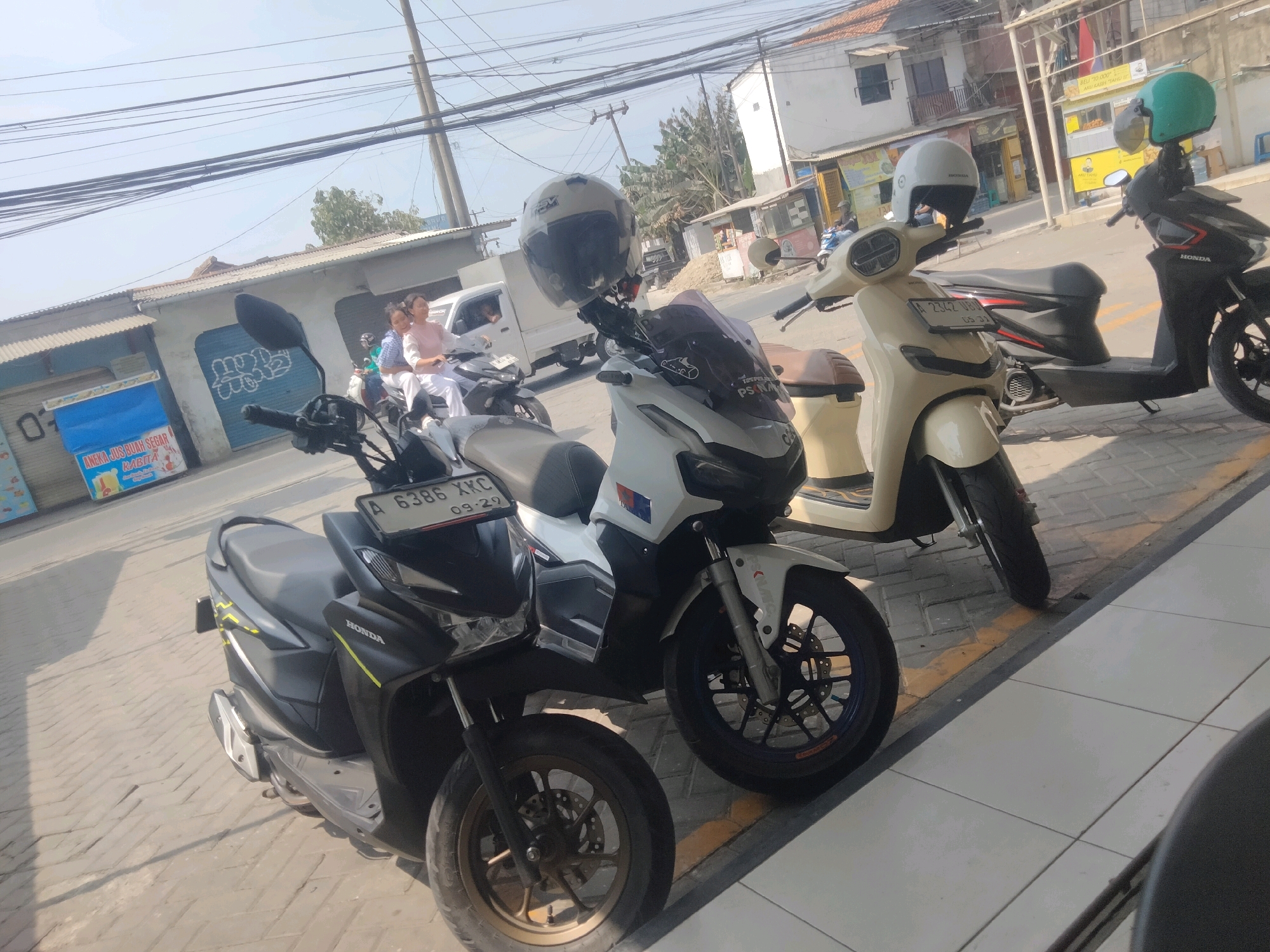 The image depicts a street scene in Pisangan Jaya, Indonesia, characterized by several parked motorbikes and pedestrian activity. In the foreground, a dark grey Honda motorbike is parked, its front wheel prominently visible. Next to it, a white scooter with a black seat is also parked, featuring a license plate that reads "A 6386 XKC" with a date of "09-29". Further to the right, a cream-colored scooter is parked, displaying a license plate "A 2342 FZC". A black motorbike with a green helmet is visible on the far right. In the background, two women are riding a motorbike, with one woman in a white top and the other in a pink top. They are passing a stall with a blue sign that says "ANEKA JUS BUAH SEGAR". The environment suggests a commercial area with buildings and overhead electrical wires. The ground is paved with interlocking concrete blocks. The lighting indicates it is daytime.