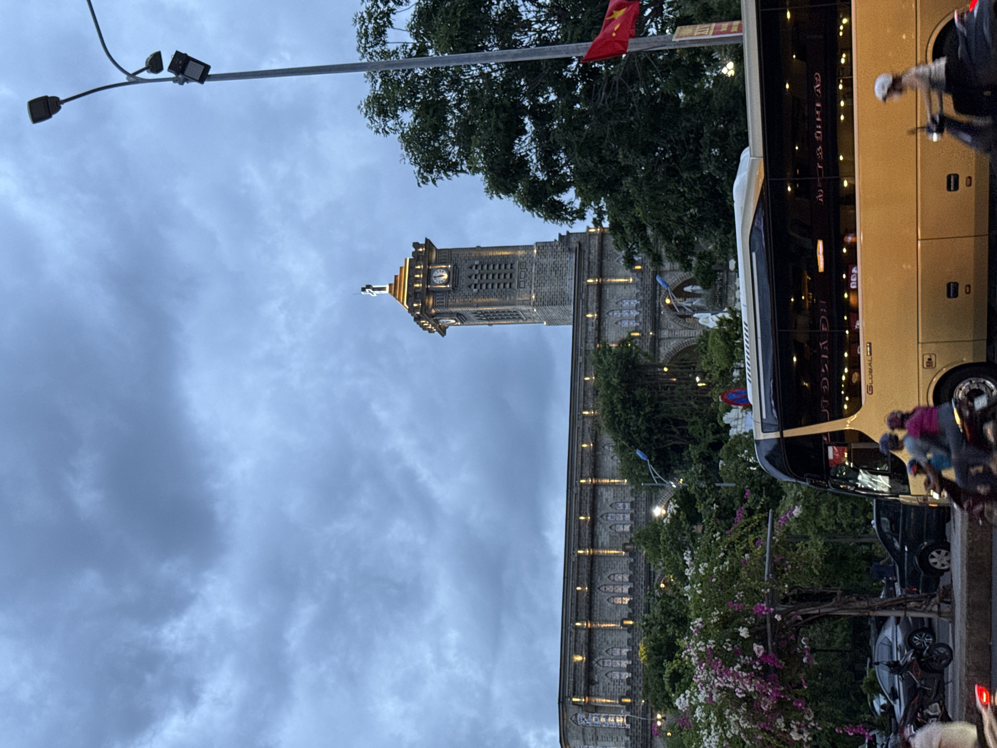 This image captures a street scene in Nha Trang, Vietnam, in the late afternoon or early evening. The prominent landmark is the Nha Trang Cathedral, a grand Gothic-style stone structure with a tall bell tower. The tower is illuminated, as are the arched windows and decorative elements along the cathedral's facade. Overcast, cloudy skies loom in the background.
In the foreground, a bright yellow tour bus is in motion, its windows reflecting the lights of the surrounding buildings. A motorbike with two passengers, a man and a woman, is also moving. Another motorbike is parked to the left, along with cars. Lush green trees and vibrant pink and white flowering bushes line the street, adding color to the urban landscape. A street light stands tall on the right side of the frame, and a Vietnamese flag is visible on a flagpole. The overall atmosphere suggests a busy city environment with a mix of historical architecture and modern transportation.