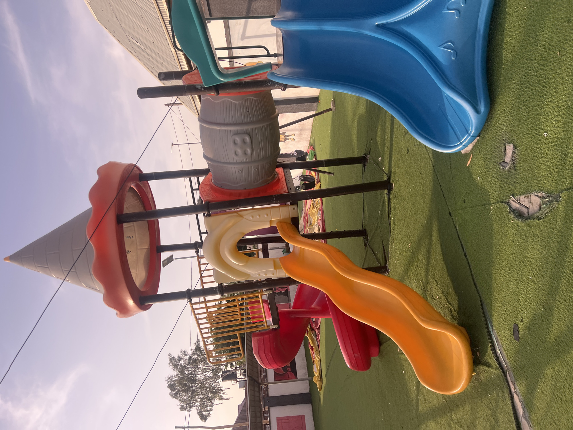 The image shows a playground with various slides and structures. The main subjects are the playground equipment, including a large blue slide, an orange slide, a yellow slide, a red slide, and a large grey barrel-like structure designed to resemble a wooden barrel. There is also an ice cream cone-shaped structure, which appears to be a part of the playground. The playground is situated on a green artificial turf surface. In the background, there is a building with a corrugated metal roof and some trees. The sky is visible, suggesting it is daytime, possibly late afternoon or early evening due to the lighting. The weather appears to be clear. There is no text visible in the image. The location is identified as Jalingo, Nigeria. The image captures a static scene of the playground, with no activity or people visible. The overall impression is of an outdoor recreational area.