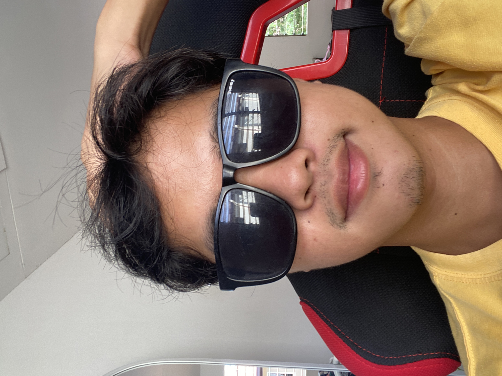 The image is a close-up, selfie-style photograph of a young person wearing black sunglasses and a yellow t-shirt. The person is seated in a black gaming chair with red accents. Their hand is raised and resting on the back of their head, with dark, somewhat messy hair visible. The sunglasses reflect a scene that appears to be indoors, with horizontal lines suggesting blinds or louvers. The environment suggests a casual indoor setting, likely a home, given the gaming chair and personal nature of the selfie. There are no visible landmarks that would definitively place this image in Davao City, Philippines. The lighting appears to be natural, coming from a window or well-lit room. The overall impression is that of a relaxed, informal moment captured by the subject.