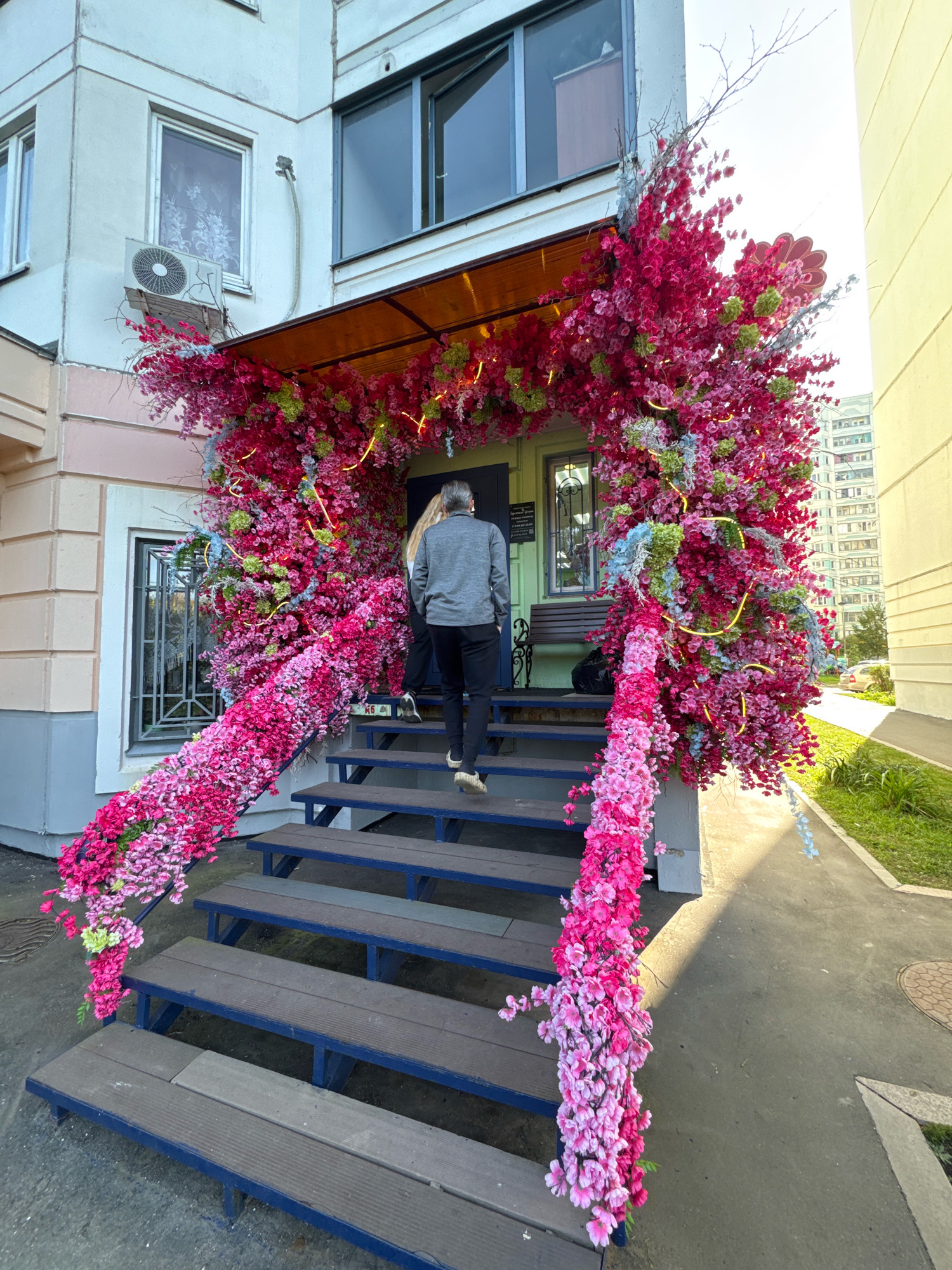 An outdoor, eye-level, medium shot captures the entrance of a building in Милицейский посёлок, Russia, adorned with an abundant artificial floral arrangement. The floral display, primarily in shades of fuchsia and pink, cascades down the sides of a set of blue-railed, wooden stairs leading to the entrance. Green and light blue accents are interspersed within the pink blooms. A person, viewed from the back and wearing a grey jacket and dark pants, is ascending the stairs, positioned on the third step from the bottom. The building facade is light-colored with visible window frames and an air conditioning unit mounted near a window. The ground surrounding the stairs is paved with concrete. To the right of the entrance, a patch of green grass and a sidewalk are visible, with a segment of a light-colored building in the background. The lighting suggests it is daytime, with shadows cast on the ground.