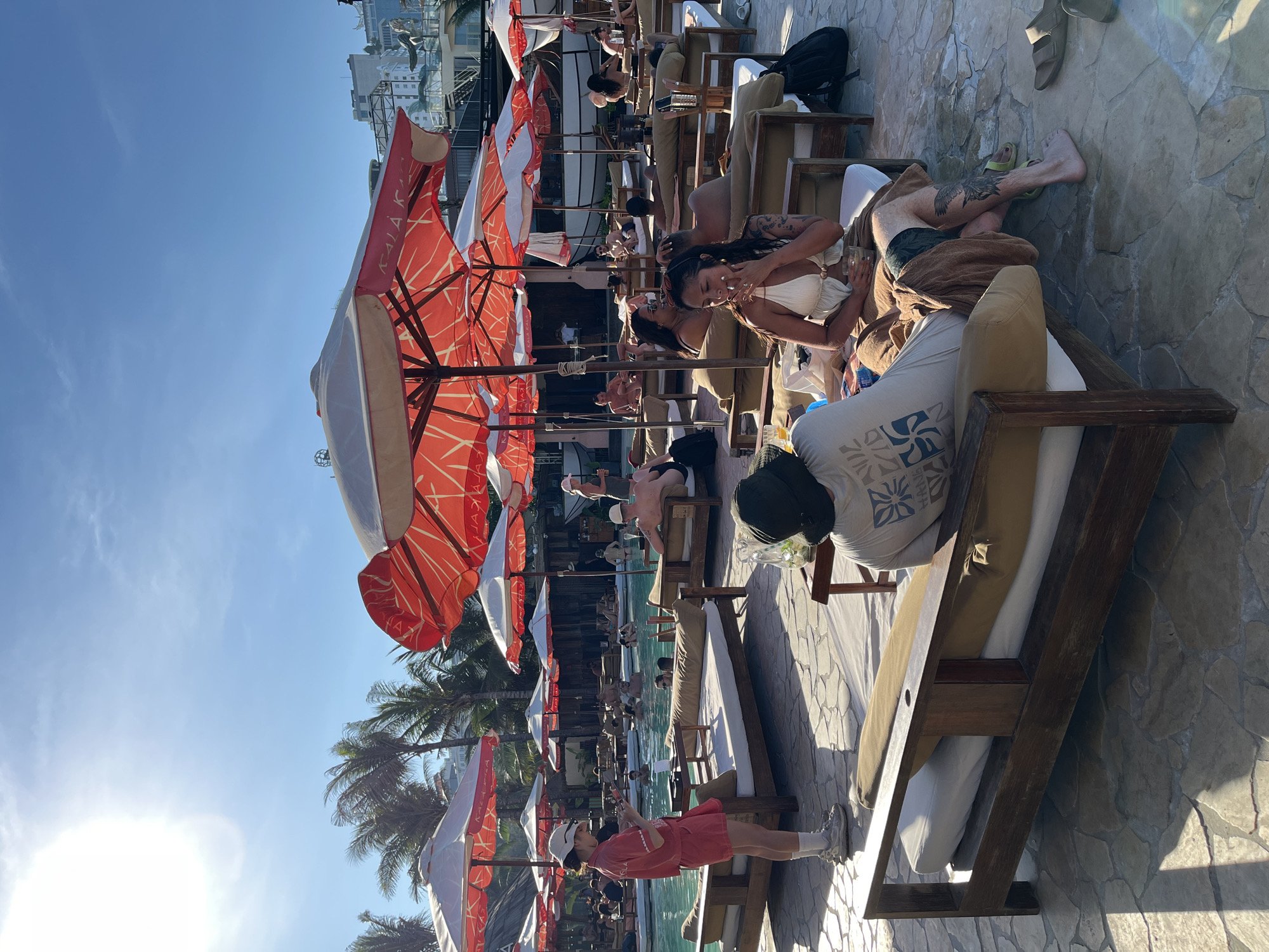 The image shows a busy outdoor pool area in Đà Nẵng, Vietnam, with numerous people relaxing on lounge chairs under red and white striped umbrellas. People are sunbathing, swimming in the pool, and socializing. Some are wearing swimwear, others casual clothing. The pool area appears to be part of a resort or hotel, with buildings visible in the background. The weather is sunny.