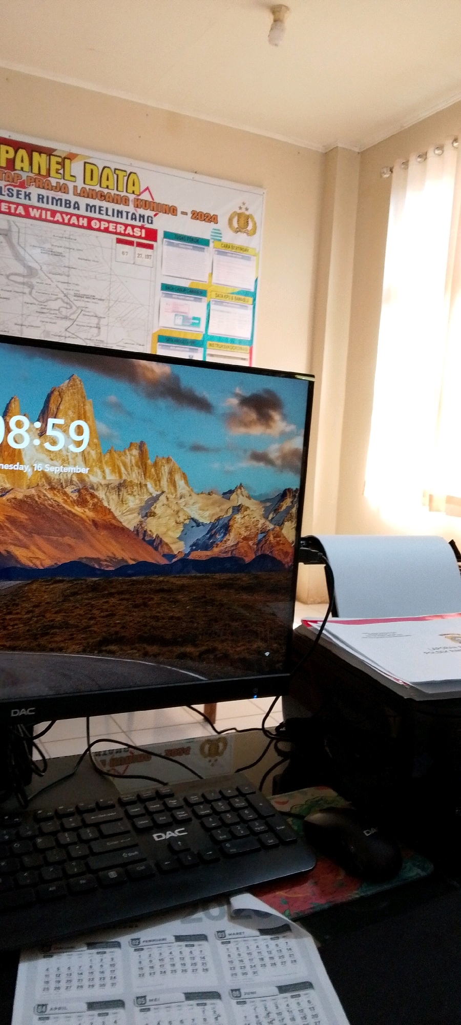 The image shows a desk with a computer monitor displaying a mountain landscape wallpaper and the time 08:59. Below the monitor, a black keyboard with the logo "DAC" is visible, along with a computer mouse. A calendar is placed in front of the keyboard, showing pages for April, May, June, February, and March. A stack of papers is to the right of the monitor. In the background, a large whiteboard with text, maps, and charts is mounted on the wall. The room appears to be an office, with a window covered by curtains to the right. The ceiling has a light fixture. The overall scene depicts a work desk setup in an office environment.