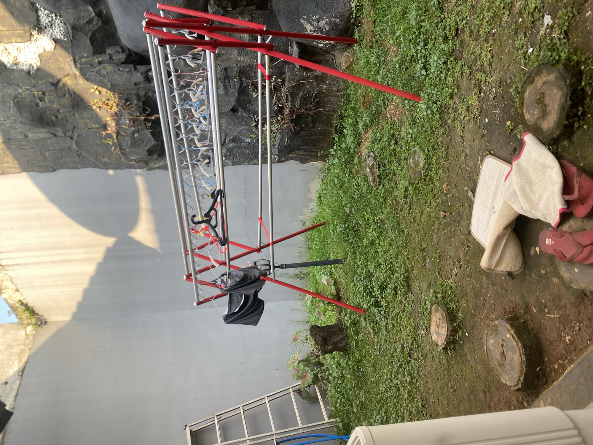 In Dukuh, Indonesia, a backyard scene unfolds under a bright sky casting sharp shadows on a gray wall. A red and silver clothes drying rack is prominent, holding several black and gray garments on hangers, along with numerous empty plastic hangers. To the left, a silver ladder leans against the wall, its blue hose coiled nearby. The ground is covered in lush green grass and scattered tree stumps, with a few plants growing near a weathered wooden stump. In the background, a rough, gray rock formation rises, dotted with small green plants. In the foreground, a rolled-up beige mat and a pair of red flip-flops lie on the ground, near a white appliance on the left.