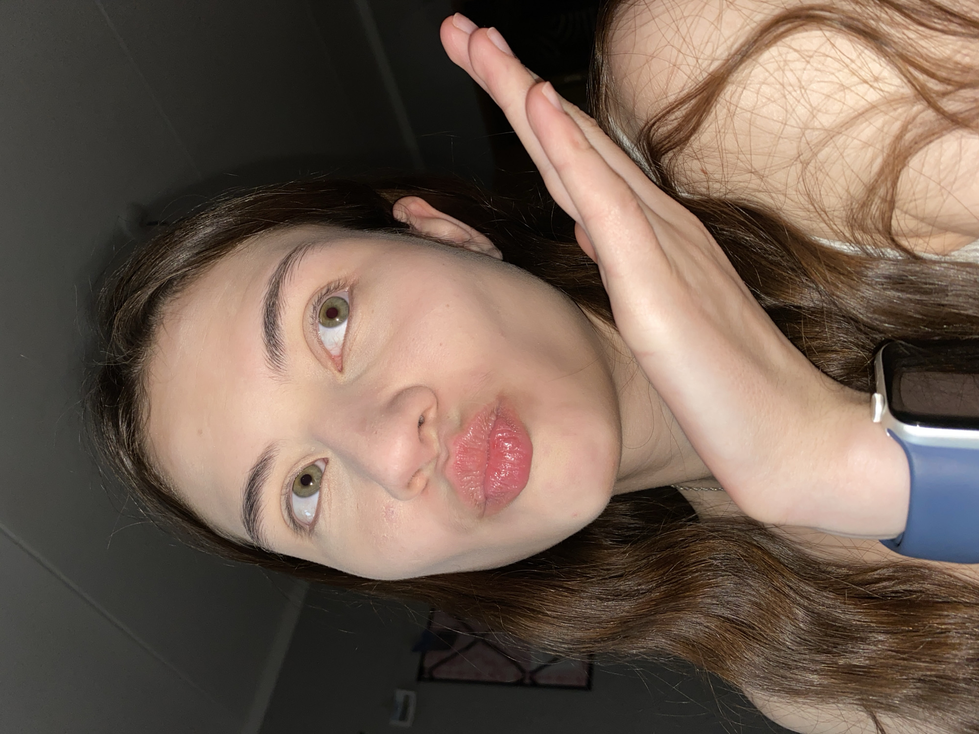 A close-up, slightly angled selfie of a young woman with light skin, long brown hair, and green eyes. She has her hand raised, palm towards the camera. She is puckering her lips in a kissing gesture. The background is dark, with a glimpse of a wall with a framed picture. The scene appears to be indoors. There is no indication of location in Votkinsk, Russia. A smartwatch with a blue band is visible on the woman's wrist.