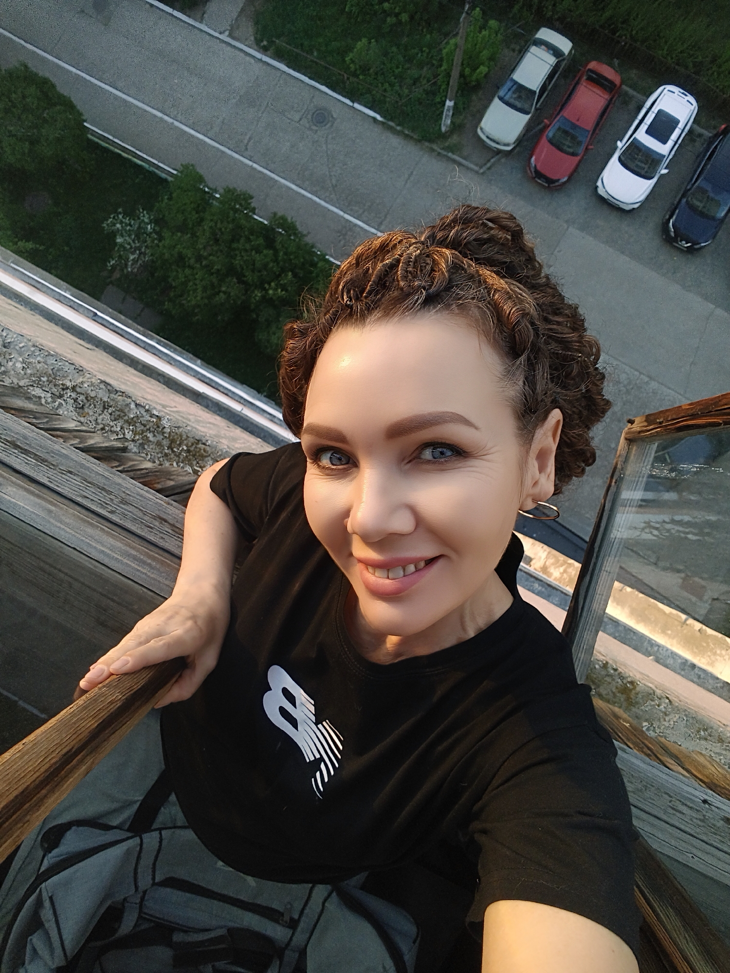 A high-angle, close-up photograph captures a woman with light skin, bright blue eyes, and curly brown hair styled in an updo. She is smiling, and her expression is friendly and inviting. She is wearing a black t-shirt with a white graphic on the chest and small hoop earrings.
The woman is positioned on what appears to be a wooden structure, possibly the top of a building or a lookout point. She is holding onto a wooden railing.
Below the railing, the scene reveals a view of a city street with parked cars. The cars, in colors of white, red, and blue, are parked neatly along the street. Beyond the cars, there are some trees and other foliage. The light suggests it is daytime, possibly late afternoon. The location in the city of Unknown, Unknown is not identifiable from the image.