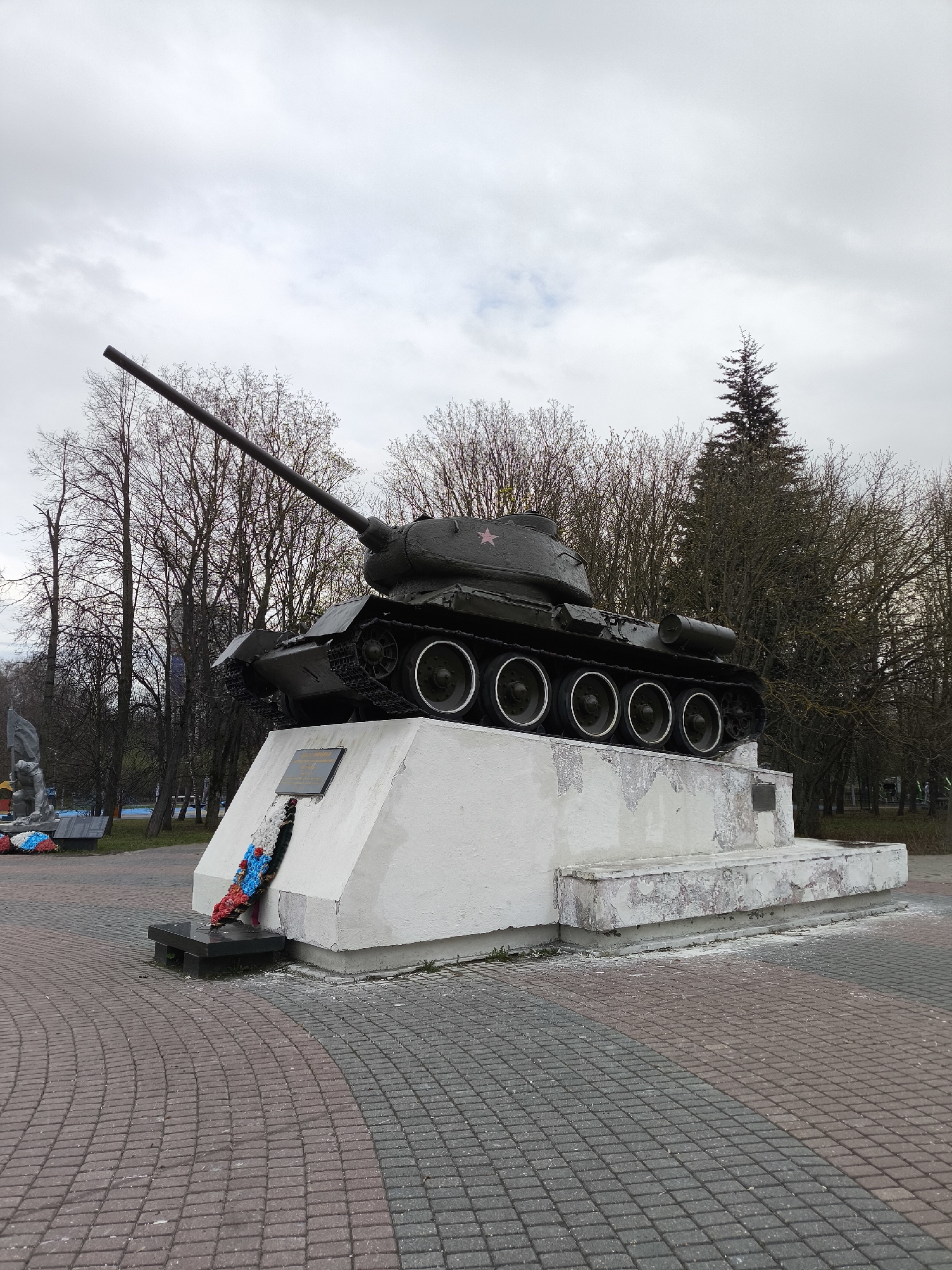 A dark green T-34 tank, typical of Soviet-era military vehicles, is prominently displayed as a memorial on a large, white concrete pedestal. The tank's long barrel points upwards and to the left, and a red star is visible on its turret. The concrete pedestal shows some signs of weathering, with patches of peeling paint or render.
At the front of the pedestal, a memorial wreath, fashioned from red, white, and blue flowers or ribbons, rests against the base, placed on a dark, low stand. Above the wreath, a rectangular dark plaque is affixed to the pedestal, but its text is not legible.
The ground surrounding the monument is paved with interlocking bricks in a pattern of reddish-brown and grey. In the background, numerous deciduous trees stand bare of leaves, suggesting either late autumn, winter, or early spring. A single, tall coniferous tree stands towards the right side of the frame, behind the tank. The sky is overcast and cloudy, indicating a daytime setting with diffused light. There are no people visible, and no discernible activity is taking place. The overall scene suggests a solemn, commemorative space, characteristic of war memorials found in public parks or squares, in this case, located in Khimki, Russia.