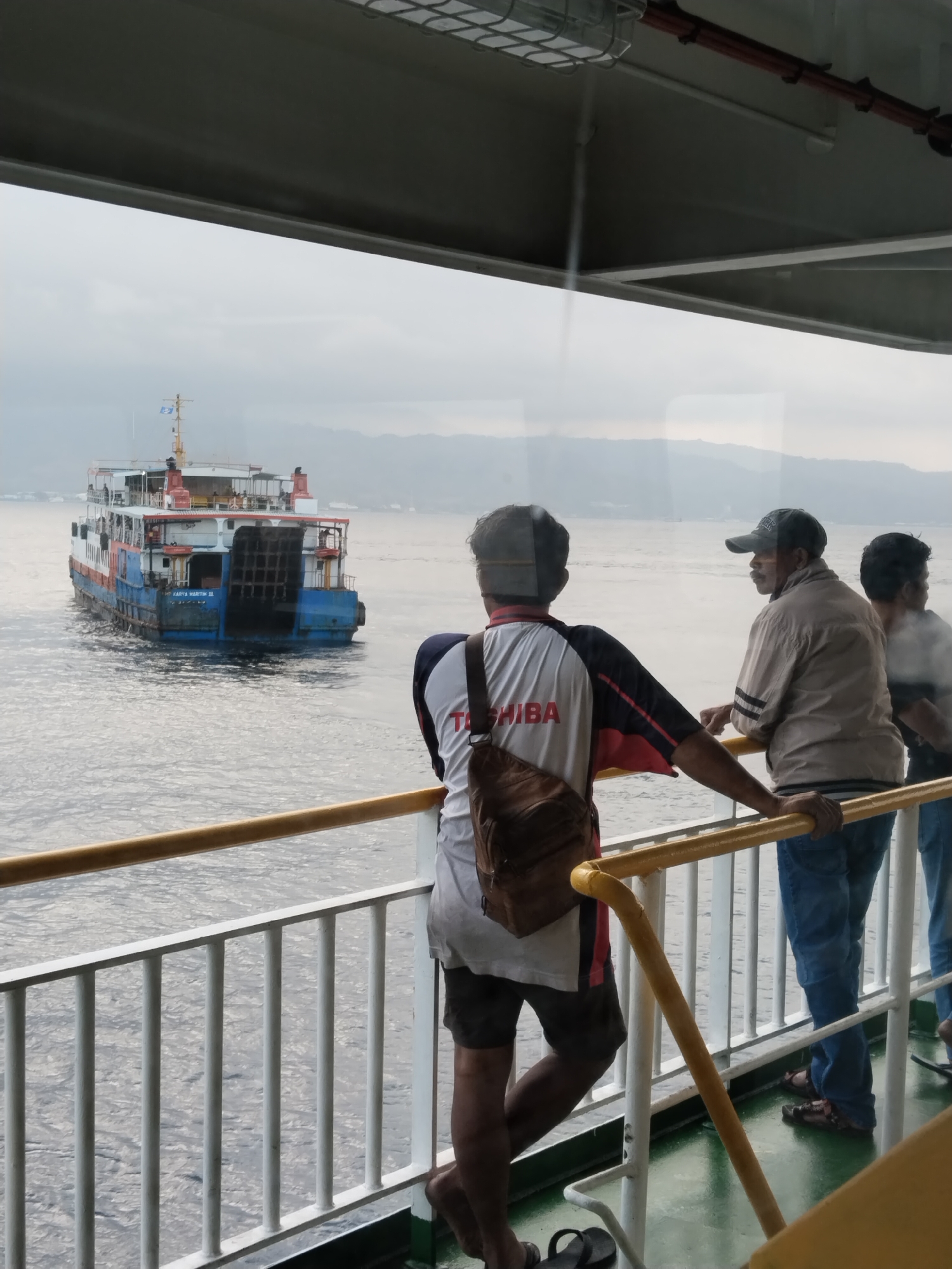 Three individuals are standing on the deck of a ferry, looking out at the ocean. A large ferry boat is visible in the distance, sailing on the water. The sky is overcast, and there are hills in the background. The scene appears to be set in Melaya, Indonesia, given the context provided. The people are casually dressed, suggesting a relaxed atmosphere. The railing and the deck of the ferry are clearly visible, indicating the viewpoint is from another vessel or a part of the same ferry.