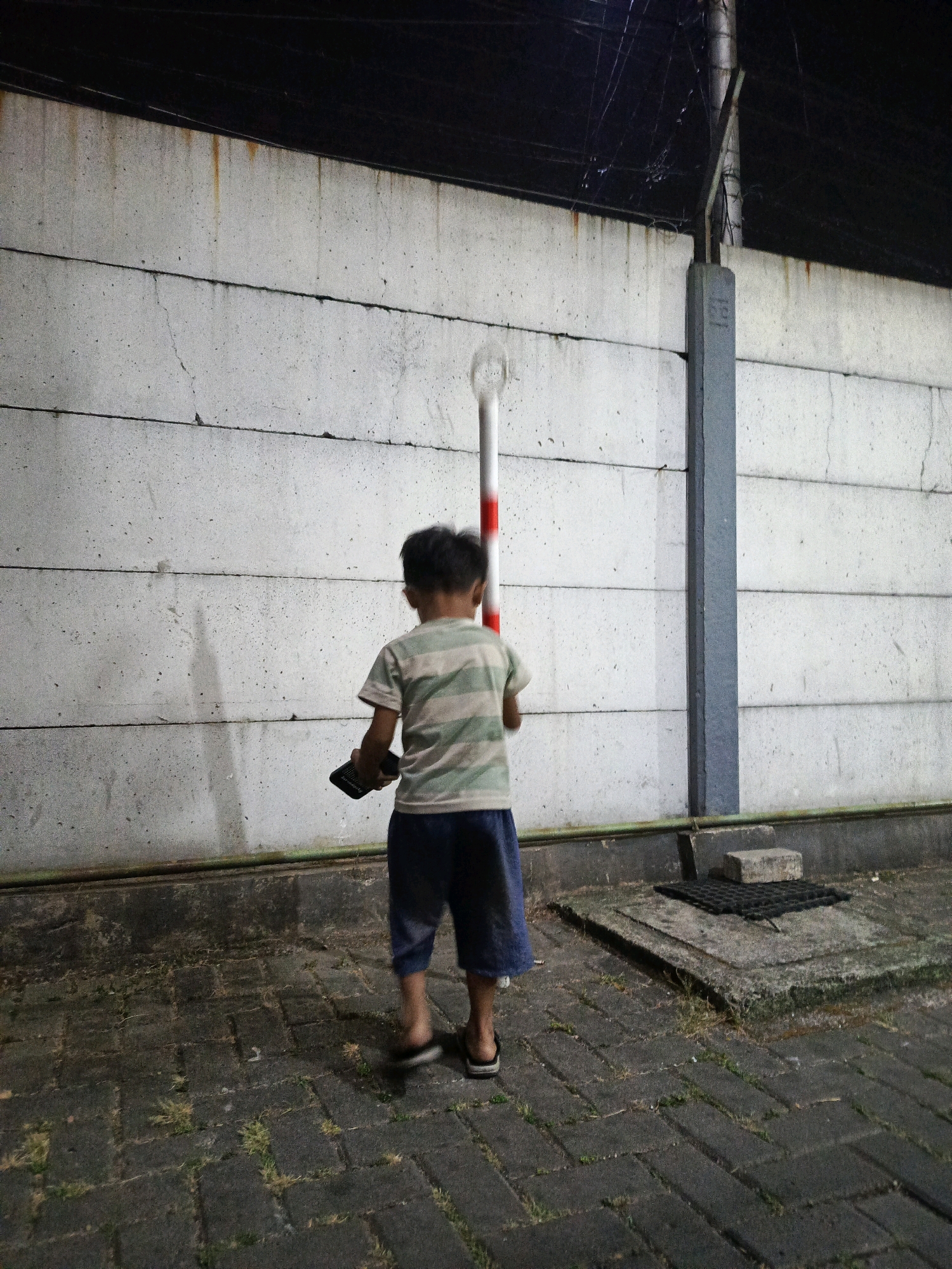 The image shows a young boy standing in front of a concrete wall at night. He is wearing a striped t-shirt and shorts and is holding a dark object in his right hand. A striped pole, red and white, is positioned behind him. The wall is made of large, horizontal concrete slabs with visible cracks and some discoloration. The ground is paved with dark, interlocking bricks, with some patches of grass growing between them. To the right, there is a concrete structure with a black grate. A grey concrete pillar is attached to the wall on the right side of the frame. The scene appears to be outdoors, possibly in a public area or alleyway, given the concrete wall and paving. The lighting suggests it is nighttime. The location is identified as East Jakarta, Indonesia.