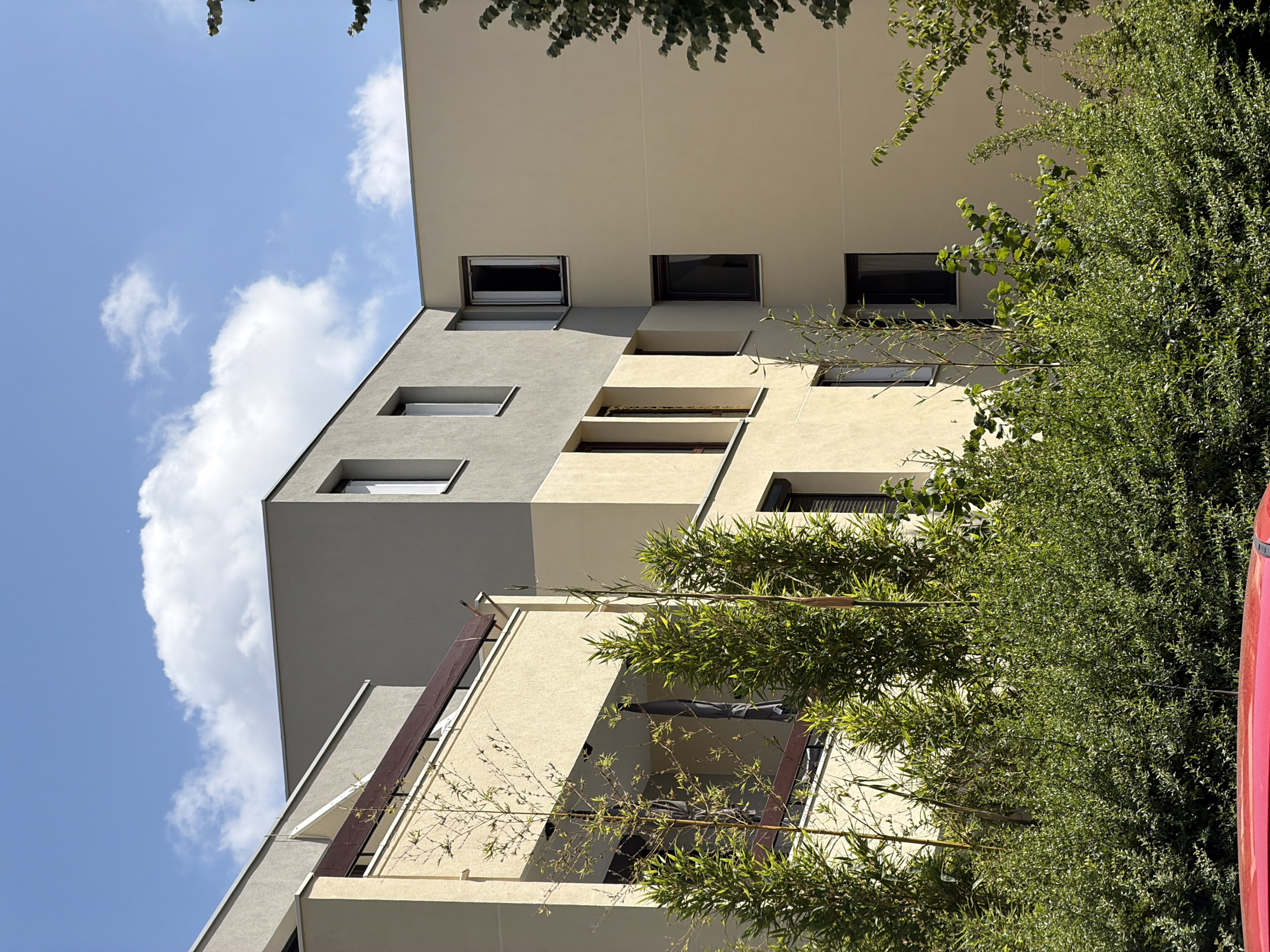 This is a low-angle shot looking up at a modern apartment building with a light beige and gray facade. The building features a series of rectangular windows, some set back into the facade, and a projecting balcony on the lower level. Lush green trees and foliage are prominent in the foreground and along the right side of the building, partially obscuring the view. The sky is a bright blue with scattered white clouds. The lighting suggests it is daytime, likely late morning or afternoon, with strong sunlight casting shadows. There is no visible text. The scene appears to be an exterior shot of a residential building in a well-maintained, possibly urban or suburban, environment.