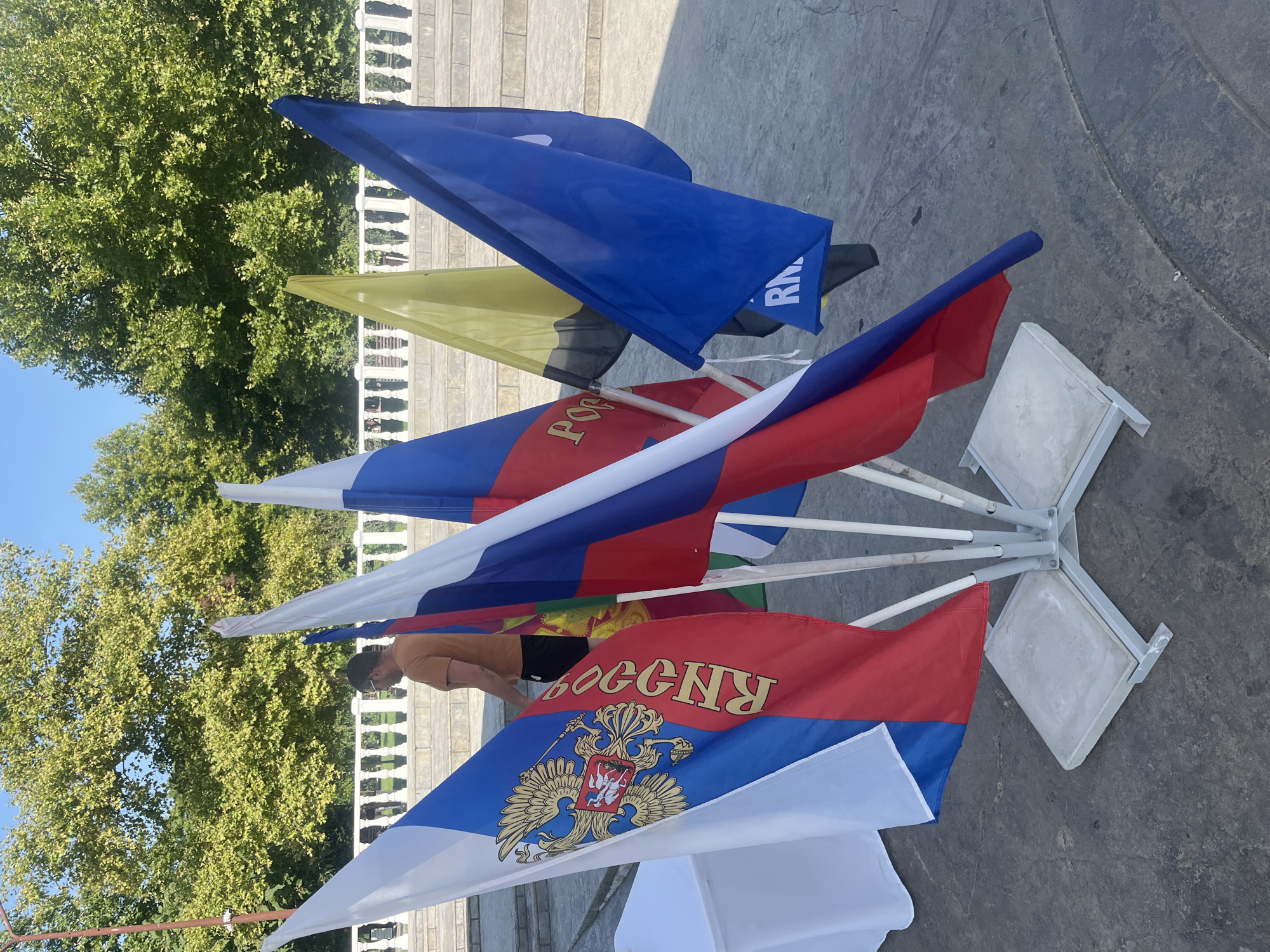A man in an orange t-shirt and dark shorts stands behind a cluster of flags on a sunny day. The flags, mounted on a metal stand with concrete bases, include the Russian flag with its coat of arms and text, a solid blue flag with white text, and a yellow and black flag. The background features lush green trees, a white balustrade, and stone steps, suggesting an outdoor public space. The ground is paved with concrete.