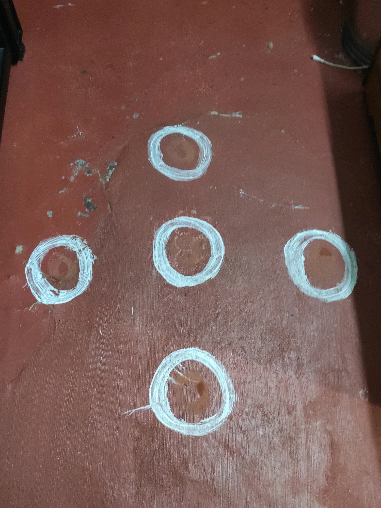 The image shows a reddish-brown concrete floor with five white circular markings painted on it. The markings are irregular and appear to be hand-drawn. One marking is at the top center, two are in the middle row, and two are in the bottom row, forming a somewhat triangular arrangement. The floor has a rough texture and some scuff marks or debris. A dark object, possibly furniture, is visible on the left side of the frame, and a shadow falls from the right. There is a thin white wire with a red tip extending from the upper right corner. The scene appears to be an indoor floor in a dwelling or common area, potentially in Hyderabad, India, given the style of floor markings sometimes seen in traditional Indian homes or public spaces.