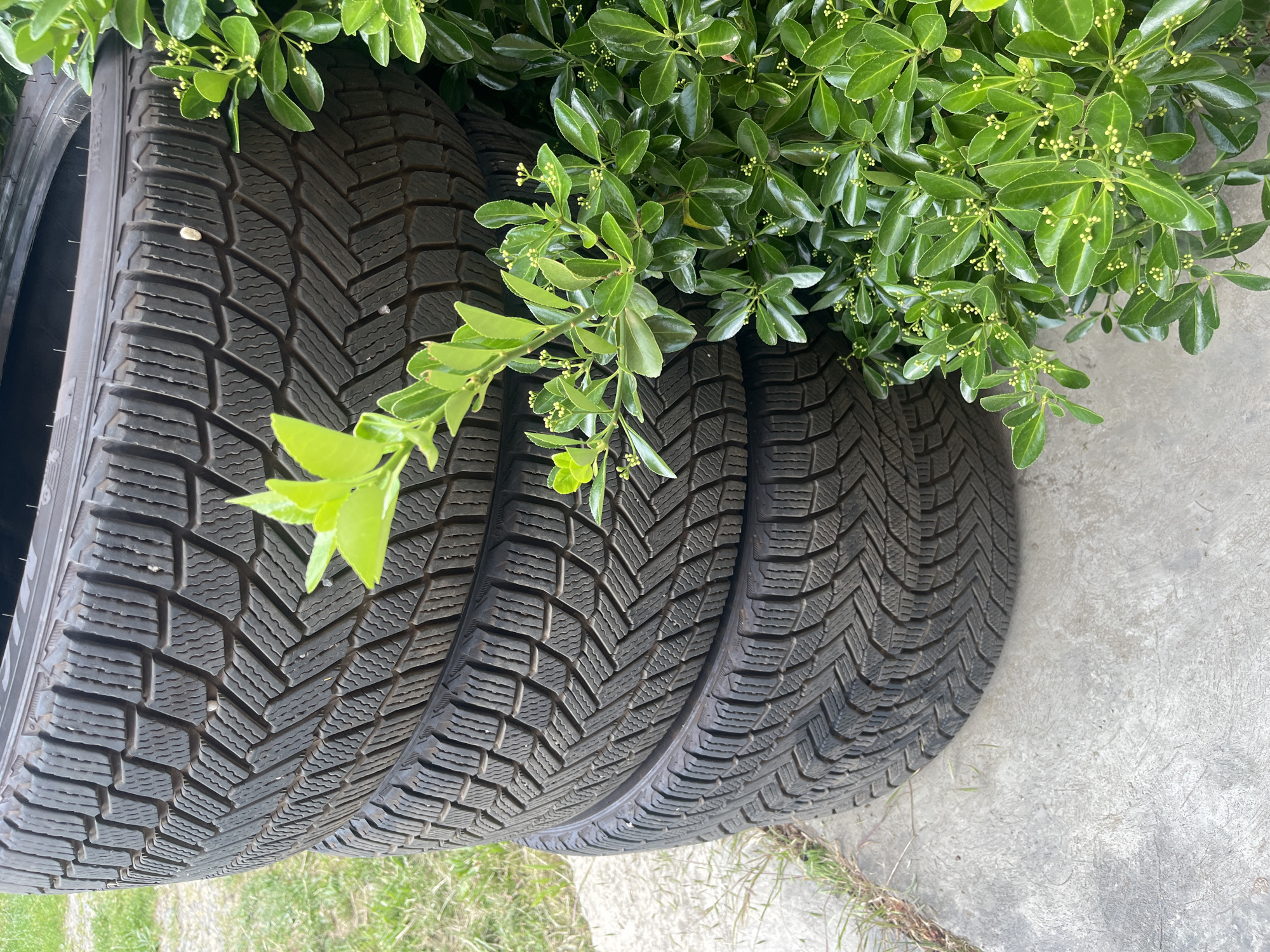 A stack of four car tires is positioned on a concrete surface next to a patch of green grass. A leafy green bush with small white buds partially obscures the tires on the right side of the image. The tires exhibit a patterned tread and are dark in color. The scene is outdoors in daylight. No people or animals are visible. The image was taken in Onokivtsi, Ukraine.