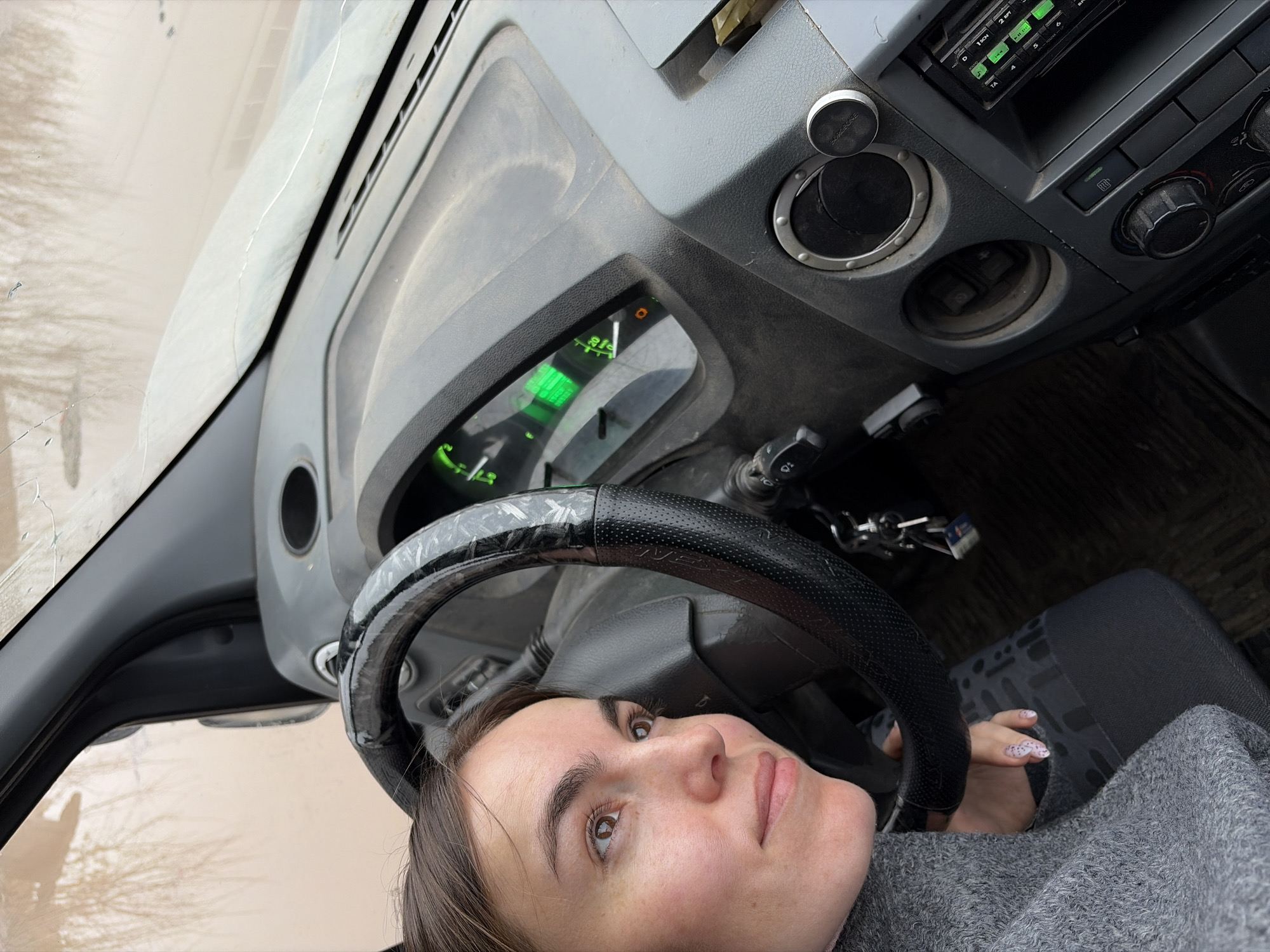A woman is positioned inside a vehicle, presumably a car, with her face turned towards the camera. The dashboard is visible, featuring various controls and a radio unit. Keys are hanging from the ignition. The exterior, visible through the windshield, appears to be an urban environment. There is no verifiable evidence to definitively confirm the location is Izhevsk, Russia.