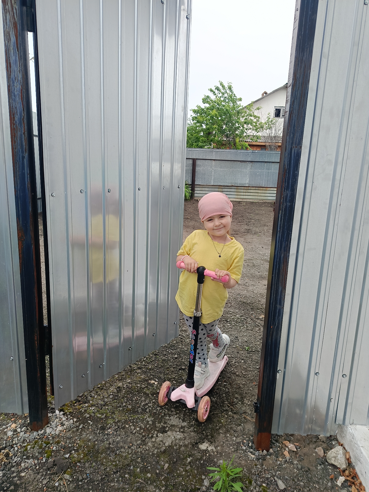 A young girl with a pink headscarf and yellow t-shirt stands on a pink scooter. She is wearing grey and white polka dot leggings and white sneakers. The girl is holding the handlebars of the scooter, which has three pink wheels. She is smiling at the camera. The background shows a corrugated metal fence with a gap, revealing a house with a green tree in front of it. The ground is covered in gravel and some small green plants. The image appears to be taken outdoors.