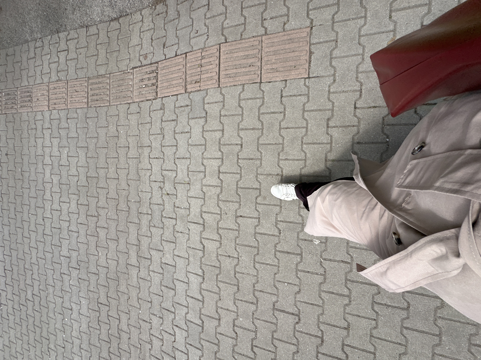 The image shows a pedestrian walkway with a tactile paving strip, indicating a pedestrian crossing or change in grade. The walkway is paved with interlocking gray concrete blocks. A person is standing on the walkway, wearing a beige trench coat and white sneakers. A red tote bag is visible. The location is likely in an urban area, with the paving suggestive of a sidewalk in a city such as Sofia, Bulgaria.