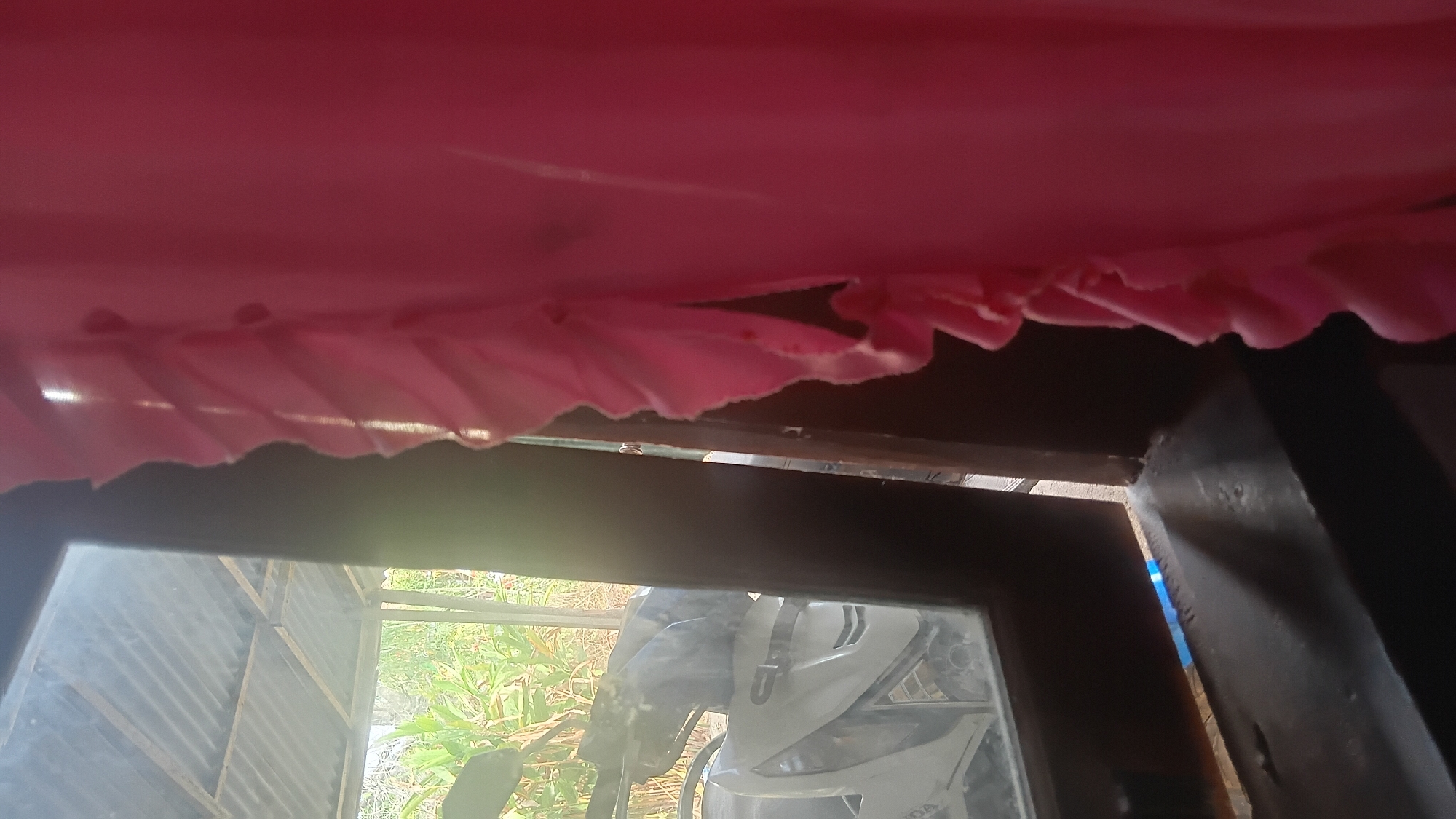 This image depicts an interior scene looking out a window in Prabumulih, Indonesia. A pink fabric with ruffled edges is prominently in the foreground, obscuring part of the view. Through the window, a white motorcycle is visible, parked under a corrugated metal roof supported by wooden beams. Green foliage can be seen in the background. The window frame appears to be dark brown wood. The overall impression is one of an everyday domestic or utilitarian space, possibly a home or workshop.