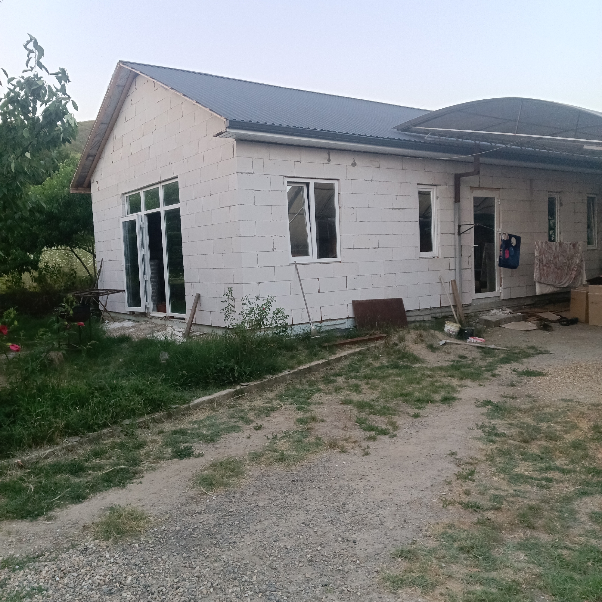 The image shows a house under construction in Prikubanskiy, Russia. The house is made of white cinder blocks and has a dark metal roof. There are several windows and a large glass door. A covered patio extends from one side of the house. The surrounding area is overgrown with grass and weeds, with some gravel and dirt paths. In the foreground, there is a small tree with red flowers and some outdoor furniture. In the background, there are more trees and a hillside. The sky is clear and pale. There are no people visible in the image.