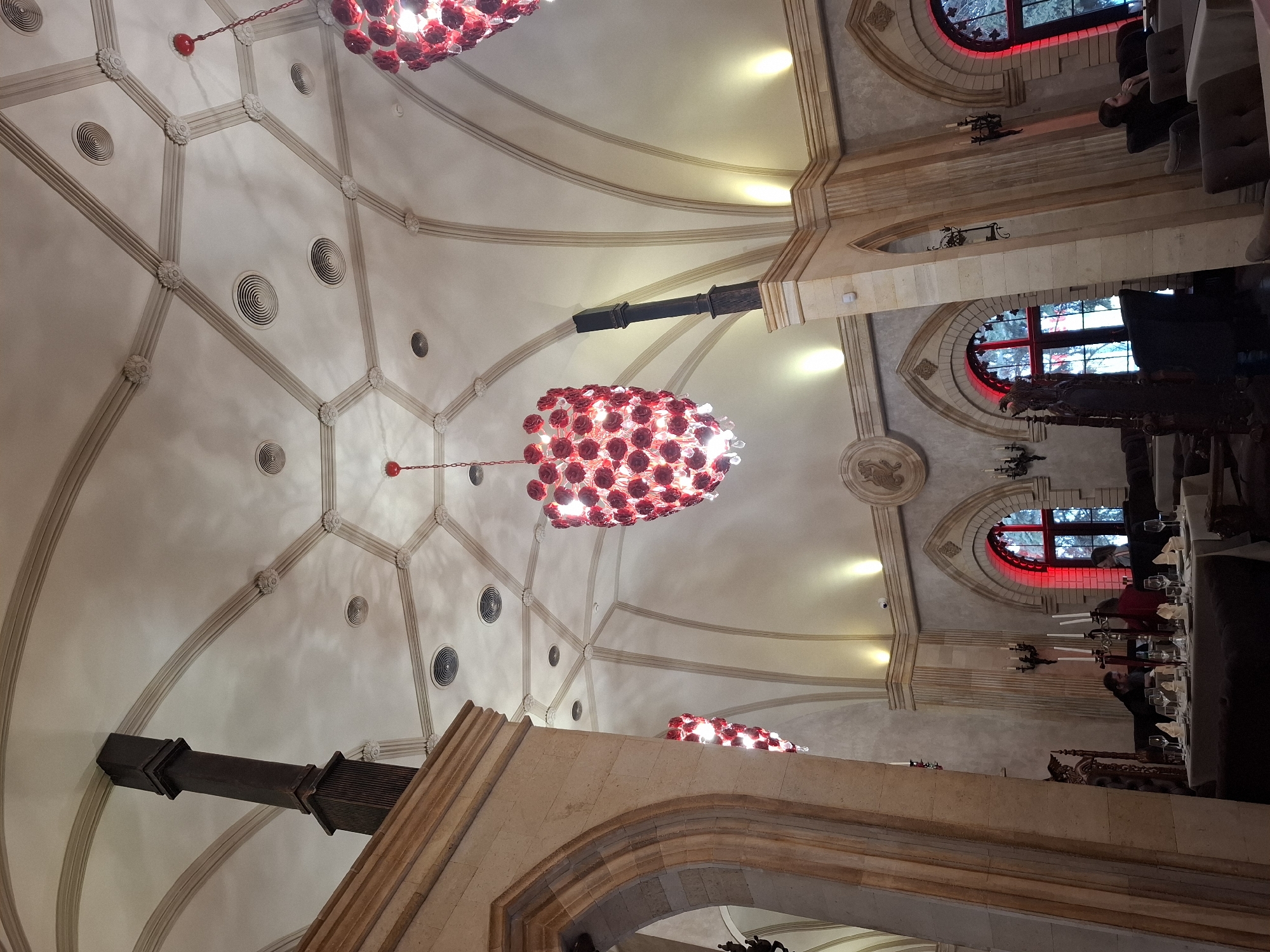 The image captures an interior view of what appears to be a dining hall or event space, potentially within a religious or historically-themed establishment. The location is inferred to be Zadonsk, Russia. The architecture features a vaulted ceiling with ornate molding and spherical chandeliers composed of red elements.
On the right side of the frame, there are arched windows with red accents. Tables are visible, appearing to be set for dining, with individuals seated. The environment has an aged, possibly gothic, style evident in the stone architecture and decorative elements.