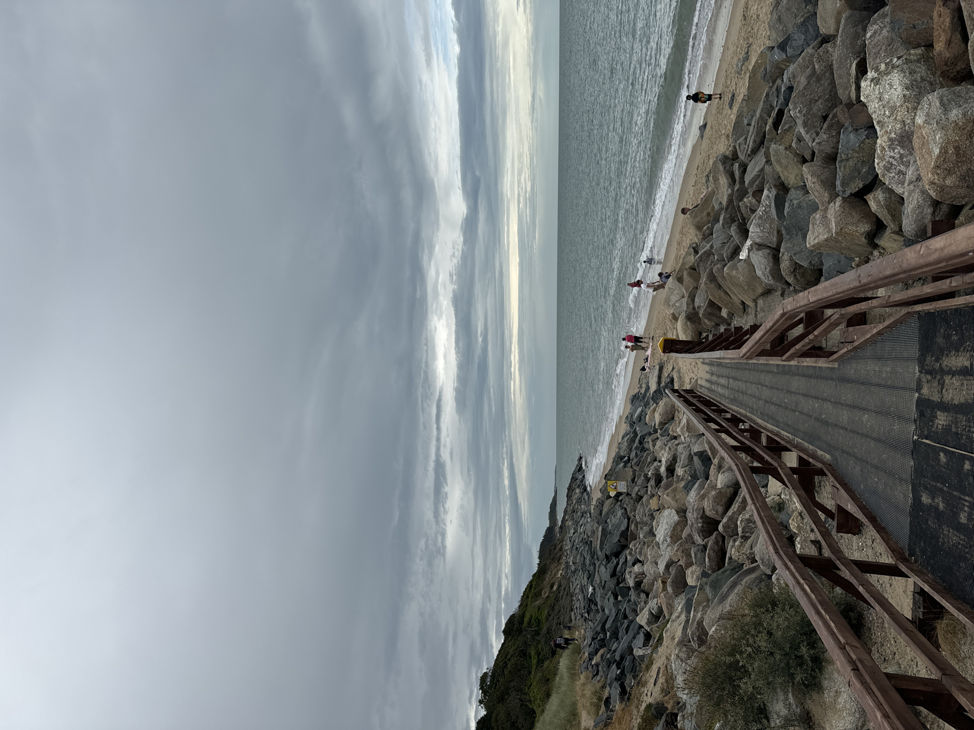 The image depicts a view of the coastline in Courtown, Ireland, under a cloudy sky. In the foreground, a wooden boardwalk with metal grating leads down towards the beach. The boardwalk is flanked by large rock formations, creating a natural barrier along the shore.
The beach itself is sandy, with gentle waves lapping at the shore. A few people are visible on the beach, some walking along the waterline and others appearing to be relaxing. To the left of the boardwalk, a grassy, vegetated slope rises, with trees and shrubs growing on its incline. The sea stretches out to the horizon, meeting the overcast sky. The overall scene is one of a serene, if somewhat gray, coastal landscape.