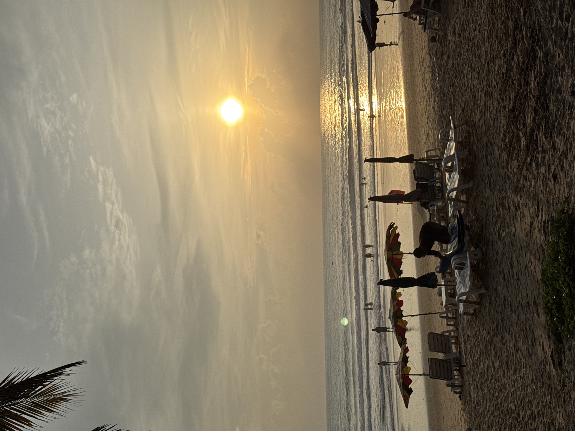 The image depicts a tranquil beach scene at sunset in Karon, Thailand. The sun, a bright orb of light, is positioned in the upper right quadrant of the sky, casting a warm, golden glow across the horizon and the ocean. The water is calm, with gentle waves rolling onto the shore. The sandy beach is lined with empty lounge chairs and colorful umbrellas, suggesting a relaxing atmosphere. In the distance, several figures can be seen wading in the shallow water, enjoying the last rays of the day. A few palm fronds frame the top left corner of the image, adding a tropical touch to the scenery. The overall mood is peaceful and serene, characteristic of a beautiful sunset on a tropical beach.