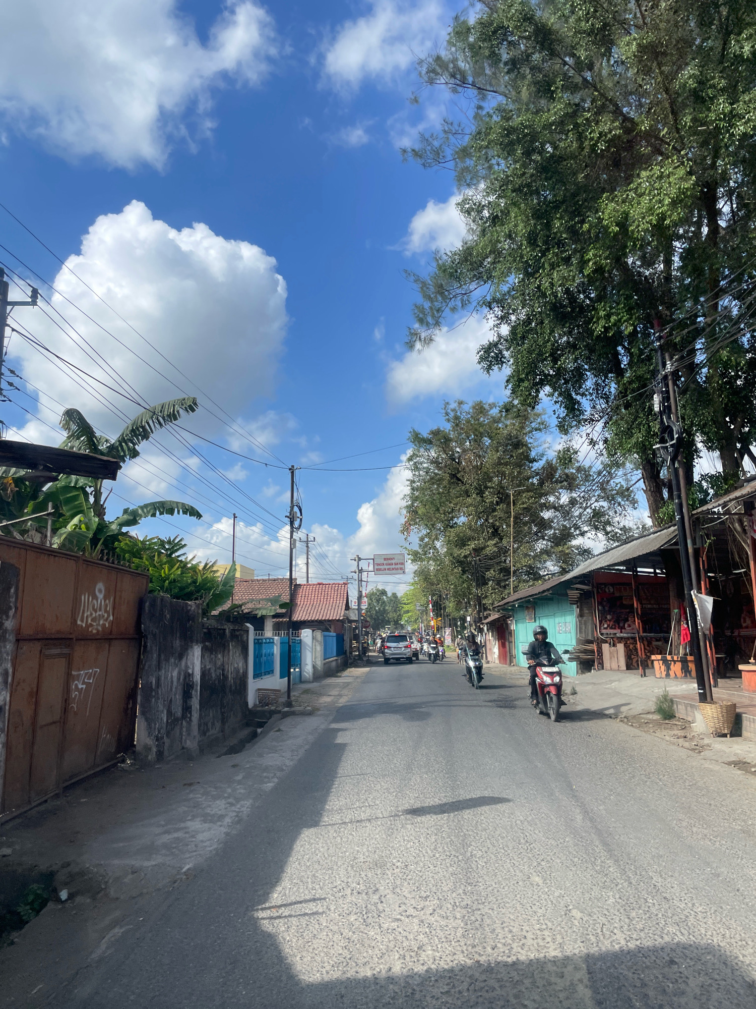 The image shows a street scene with a blue sky filled with white clouds. On the left side of the road, there is a rusty brown gate with graffiti. Adjacent to the gate is a concrete wall and some lush green vegetation, including banana trees. Further down the street, there are several buildings with red-tiled roofs and blue accents.
On the right side of the road, there are more buildings, some of which appear to be shops or stalls with awnings. Large trees with green foliage are visible, casting shadows on the road.
Several people are on the road, mostly on motorcycles. One person is riding a red motorcycle in the foreground, wearing a helmet and dark clothing. Other motorcycles and a car are visible further down the street. There are also power lines and utility poles throughout the scene. The road surface is asphalt with a textured pattern. A signpost with text in Indonesian is visible in the distance. The overall impression is of a busy street in a tropical or subtropical urban environment on a sunny day.