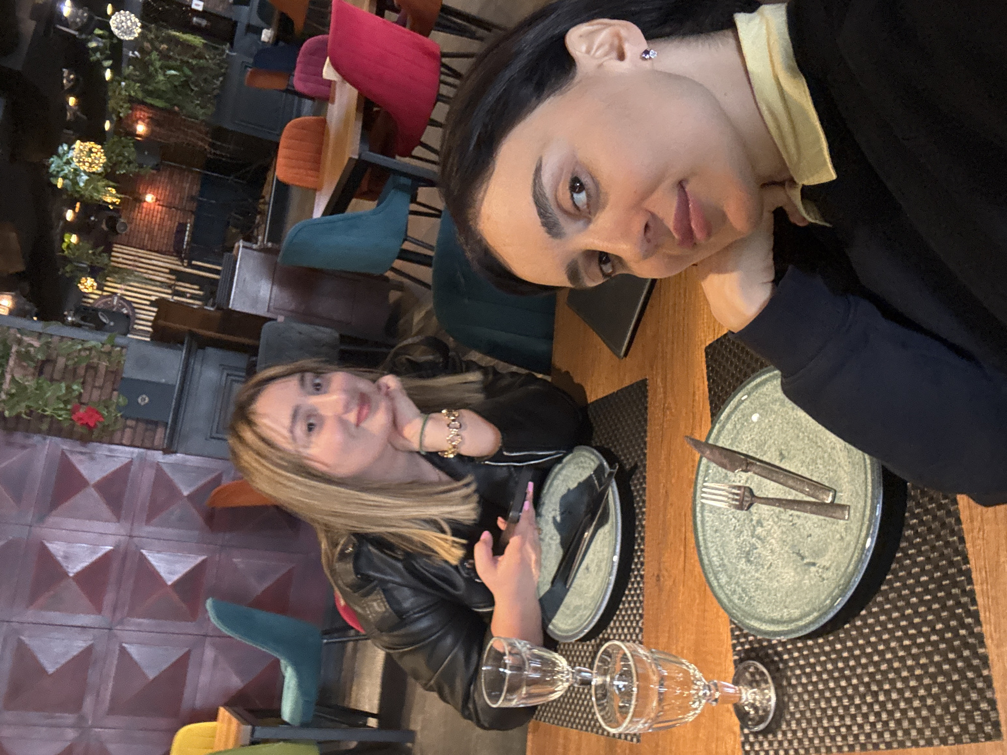 Two women are seated at a table in a restaurant, likely located in the city of Tbilisi, Georgia. One woman has long dark hair and is wearing a black top with a yellow collar. The other woman has shoulder-length blonde hair and is wearing a black leather jacket. They are sitting at a wooden table set with plates, cutlery, and glasses. The restaurant has a varied seating arrangement with chairs of different colors. The decor includes what appears to be a patterned wall and greenery, with some lights illuminating the background.