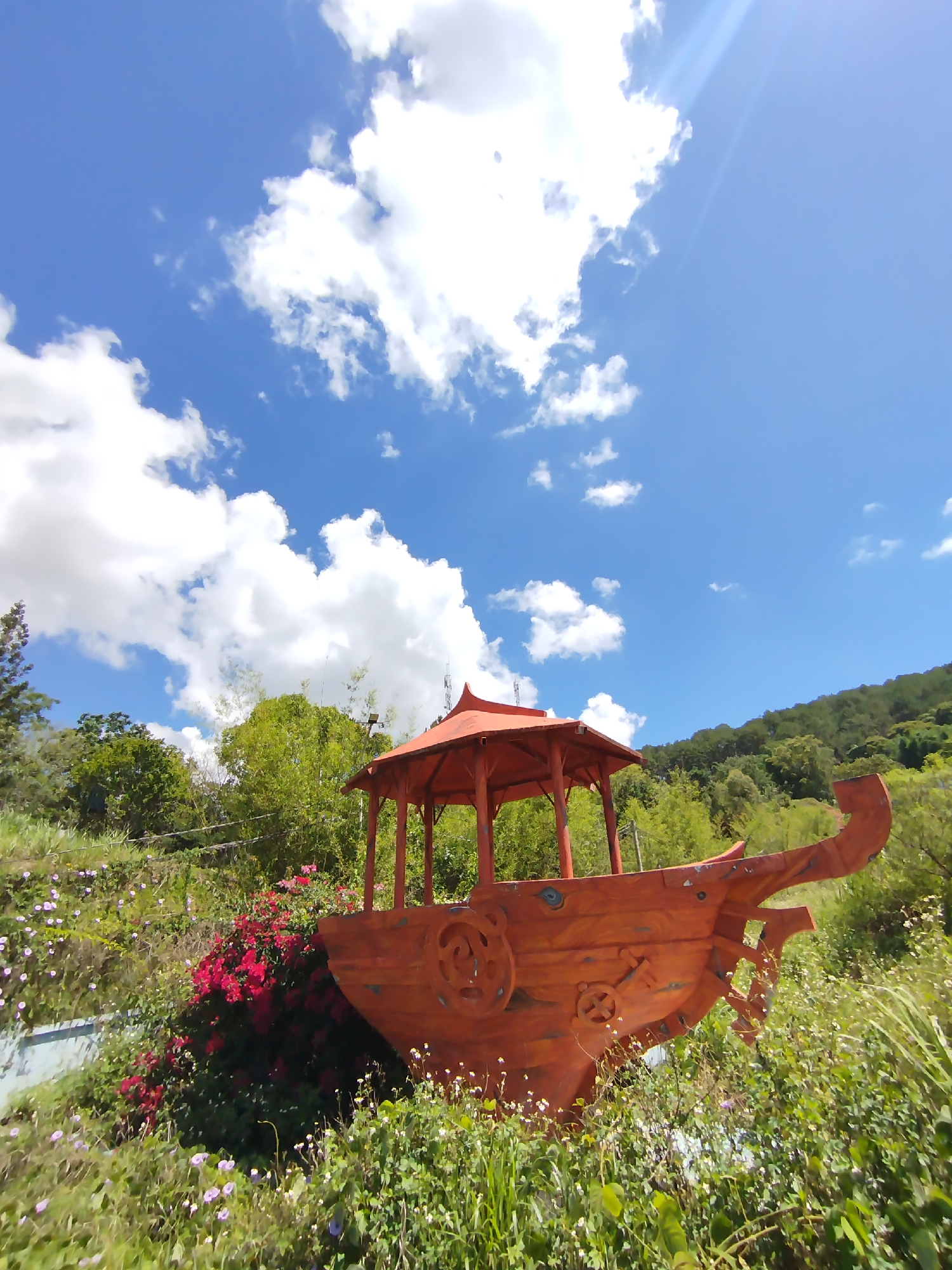 The image shows a decorative boat structure, likely a tourist attraction or whimsical garden feature, set amidst lush vegetation in what appears to be a hilly or mountainous area. The boat is made of wood, painted orange, and has a small gazebo-like structure on top. The surrounding greenery includes a variety of flowers and foliage, with a hillside covered in trees visible in the background. The sky is blue with scattered white clouds, and the lighting suggests it is a sunny day. The location is assumed to be in the city of Da Lat, Vietnam.