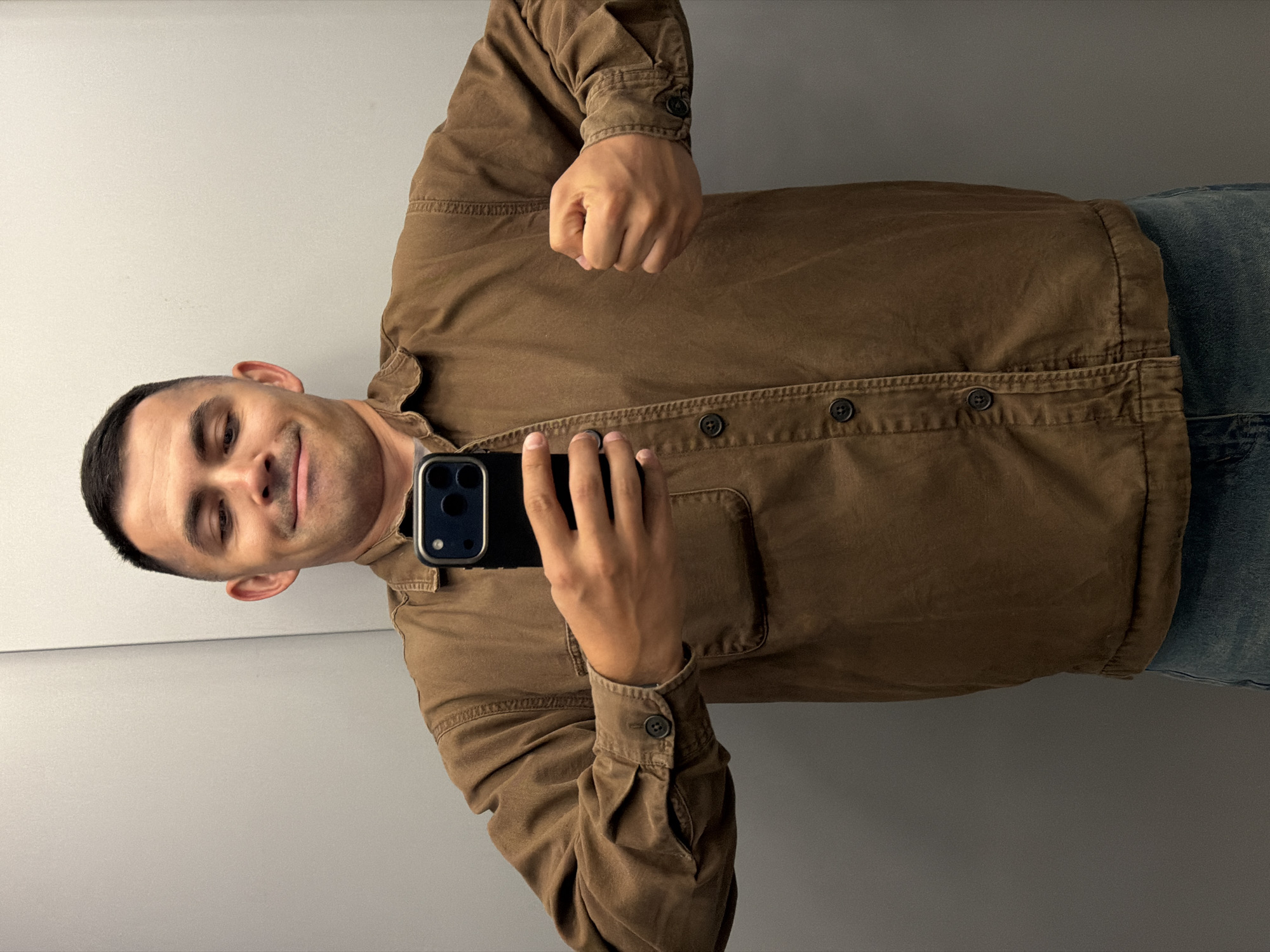 A man is captured in a mirror selfie, originally oriented 90 degrees counter-clockwise, showing him from the waist up against a plain light grey wall in an indoor setting in Izhevsk, Russia. He holds a black smartphone with a triple-lens camera system in his left hand, pointing it towards the mirror to take the photograph. His right hand is clenched into a fist, raised slightly in front of his chest. He has short, dark hair and is looking at his reflection with a subtle smile or smirk, conveying a sense of confidence or playfulness. He is wearing a long-sleeved, collared brown button-up shirt with a chest pocket on the left side and light blue denim jeans. The shirt has visible stitching details and dark buttons. The indoor lighting is even and bright. No text is visible in the image.