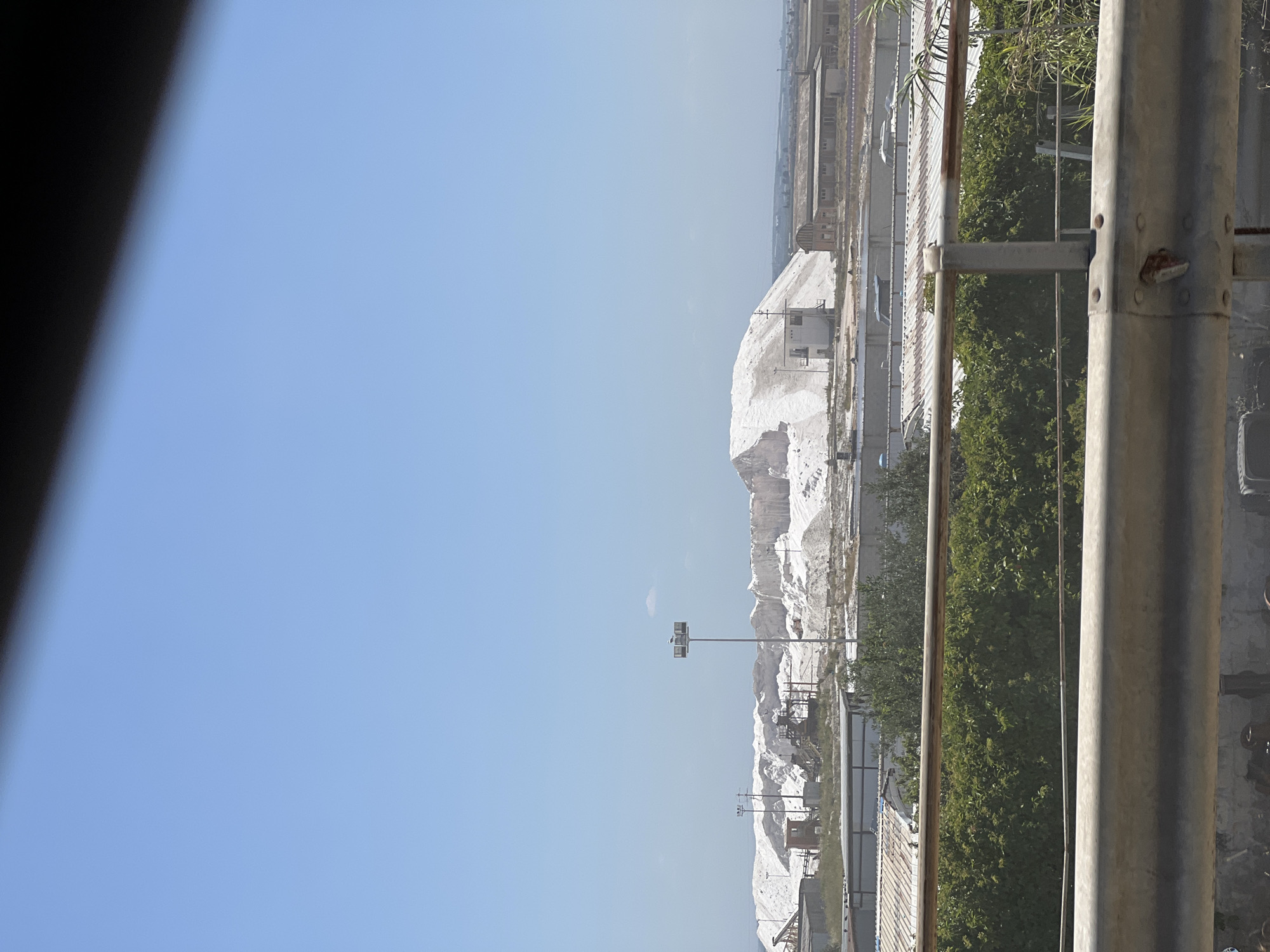 The image depicts a vast expanse of white salt mountains, characteristic of the salt pans in Margherita di Savoia, Italy. The salt mountains are a prominent feature, illuminated by a bright, clear sky. In the foreground, there are industrial structures, including buildings with corrugated metal roofs and what appear to be processing or storage facilities. Several tall light poles with lamps are visible among the salt piles. The landscape is arid, with sparse vegetation in some areas. A metal railing runs along the bottom of the frame, suggesting the photo was taken from an elevated position, possibly a bridge or an overpass. The overall scene conveys an industrial and operational environment related to salt extraction and processing.