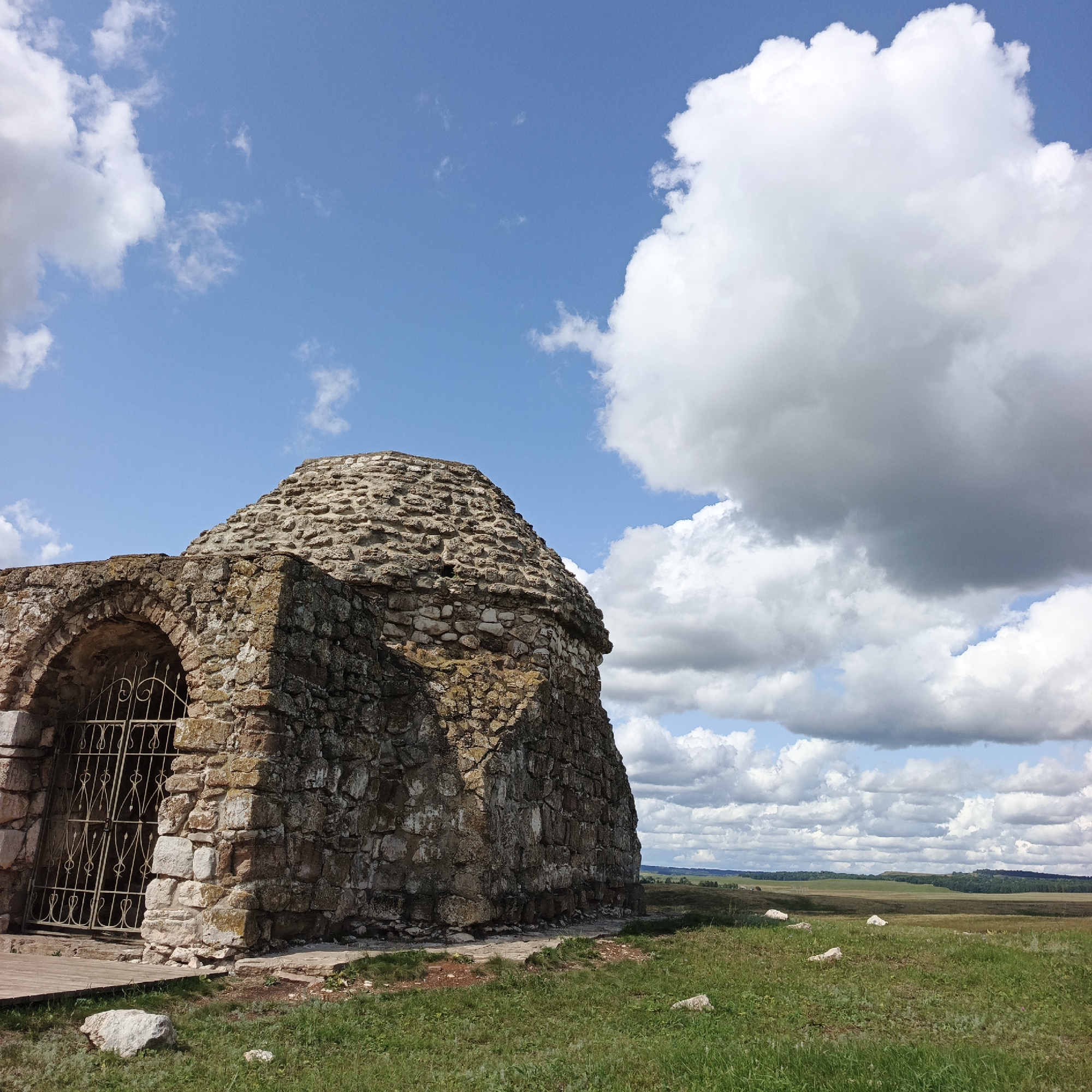 An ancient stone structure, likely a mausoleum, stands in a grassy field under a cloudy blue sky. The structure is made of rough-hewn stones and has a rounded, conical roof and an arched entrance with a decorative metal gate. The surrounding landscape is open and hilly, with a line of trees in the distance. The image was taken in Verkhnie Termy, Russia.
