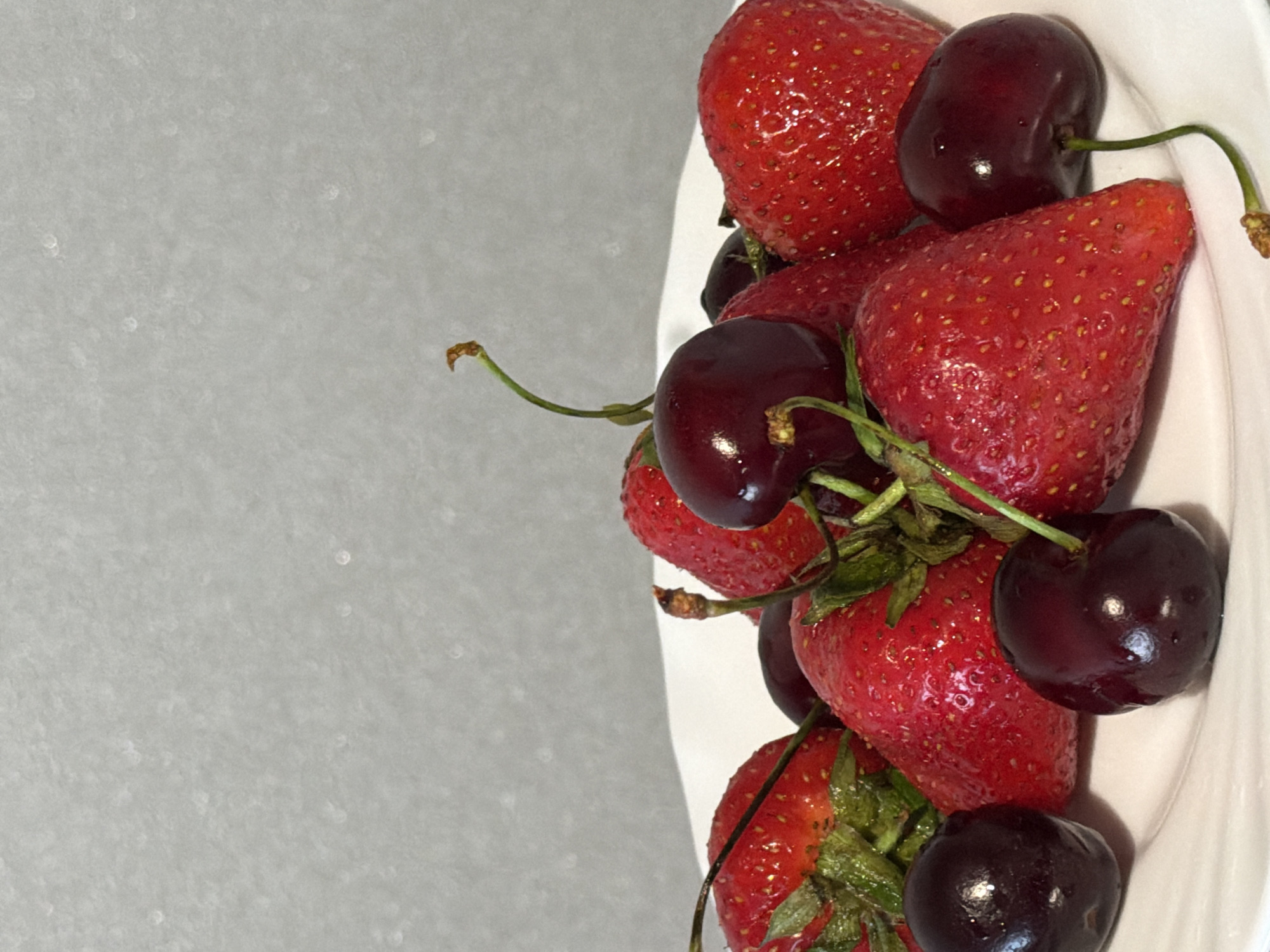 A close-up, vertical shot displays a collection of ripe strawberries and dark cherries arranged on a white plate. The fruits are clustered together, with some stems still attached to the cherries. The background is a textured, light gray surface with subtle speckles. The lighting is soft, creating a gentle sheen on the glossy fruit surfaces. No people are visible in the scene. The location is Unknown, Unknown.