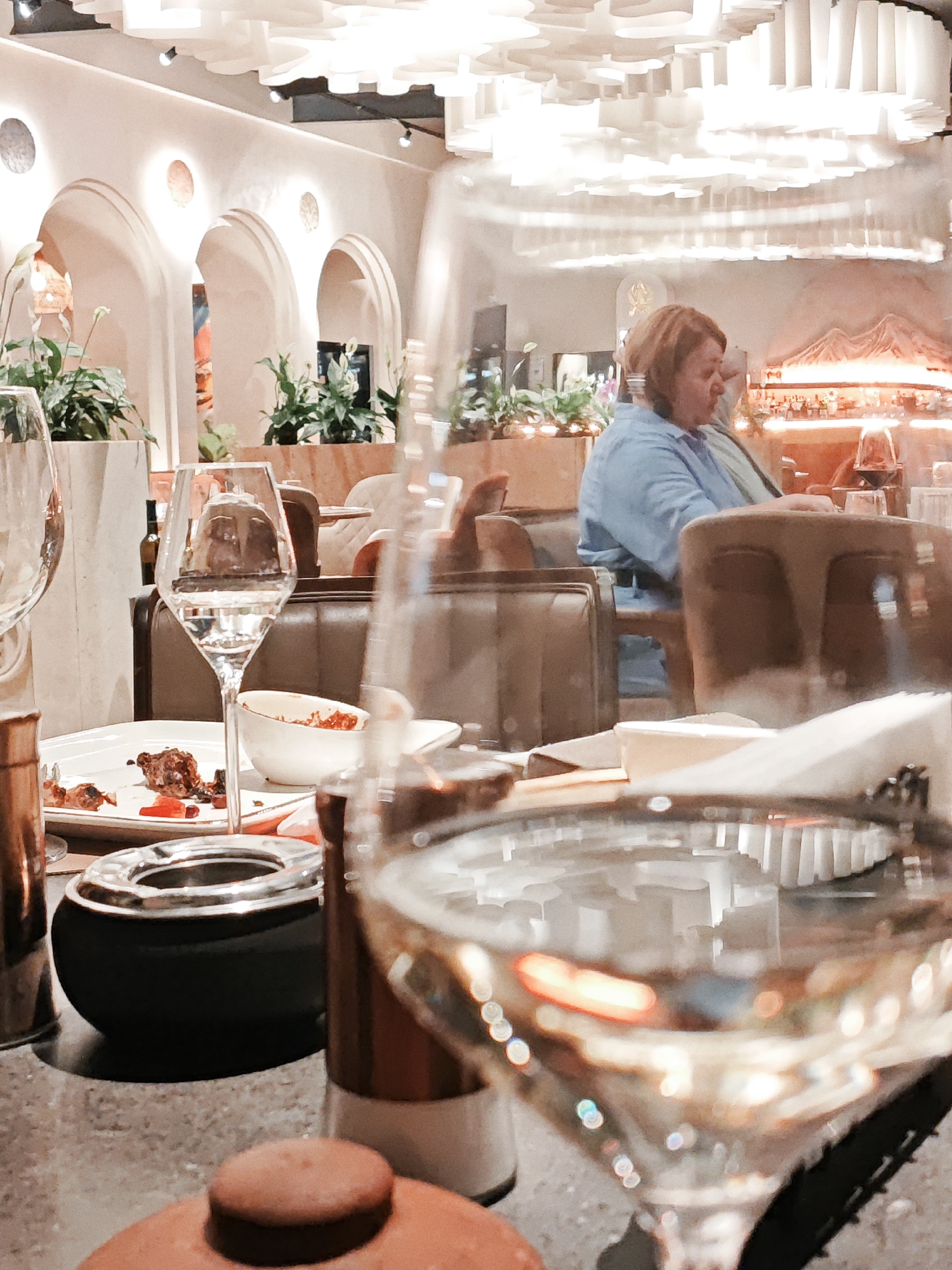 An indoor scene depicts a dining establishment with soft lighting. In the foreground, a glass of white wine is prominently featured, with reflections of the restaurant's interior visible within it. To the left, a white plate with remnants of food and cutlery rests on the table, accompanied by a white bowl containing food. An ashtray is situated between the plate and the glass. In the mid-ground, a woman wearing a light blue top is seated at a table, facing away from the camera and towards a bar area, where bottles of alcohol are displayed. Behind the woman, plants are visible in decorative planters. The background reveals an architectural design with arches and a decorative ceiling. The overall ambiance suggests a relaxed dining experience.