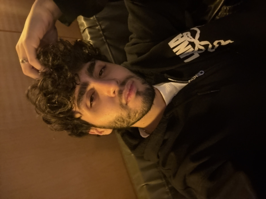 A forensic description of the scene reveals a young adult male, seated indoors in Yerevan, Armenia. The individual possesses a light-to-medium skin tone, dark curly hair, and a dark beard and mustache. He wears a black zip-up hoodie over a white undershirt, with a silver-colored chain visible beneath the shirt collar. A partial logo, possibly "LANAI" with a coiled snake graphic, is present on the right sleeve of the hoodie. His right hand is raised, with fingers interacting with his hair, and a ring is visible on his right ring finger. He maintains direct eye contact with the camera. The background consists of a plain, light brown wall or paneling on the left, and a darker, less defined area on the right, suggesting an upholstered backrest or similar structure. The lighting is dim and warm, illuminating the subject from the front-left.