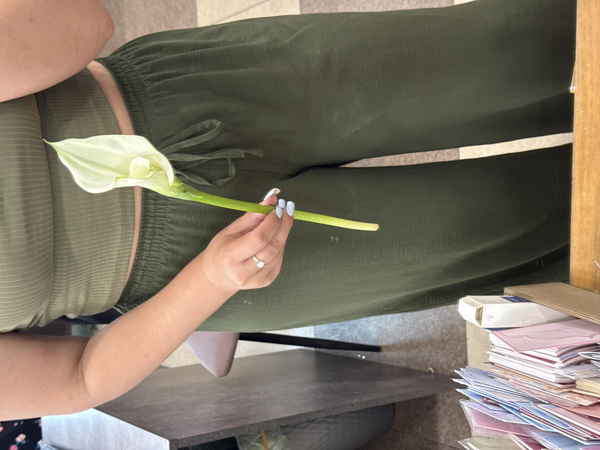 A person, partially visible from the chest down, stands holding a single white Calla Lily. They are wearing a ribbed olive green crop top and loose olive green pants with a drawstring waist. Their left hand, adorned with a diamond ring and light blue nail polish, is supporting the flower's stem. In the foreground, a wooden box filled with colorful greeting cards is visible. The background shows a dark grey surface that appears to be a desk or counter, with a portion of a brown chair and a dark object resembling a trash can. The lighting is natural and appears to be indoors. The image was taken in Assinovskaya, Russia.