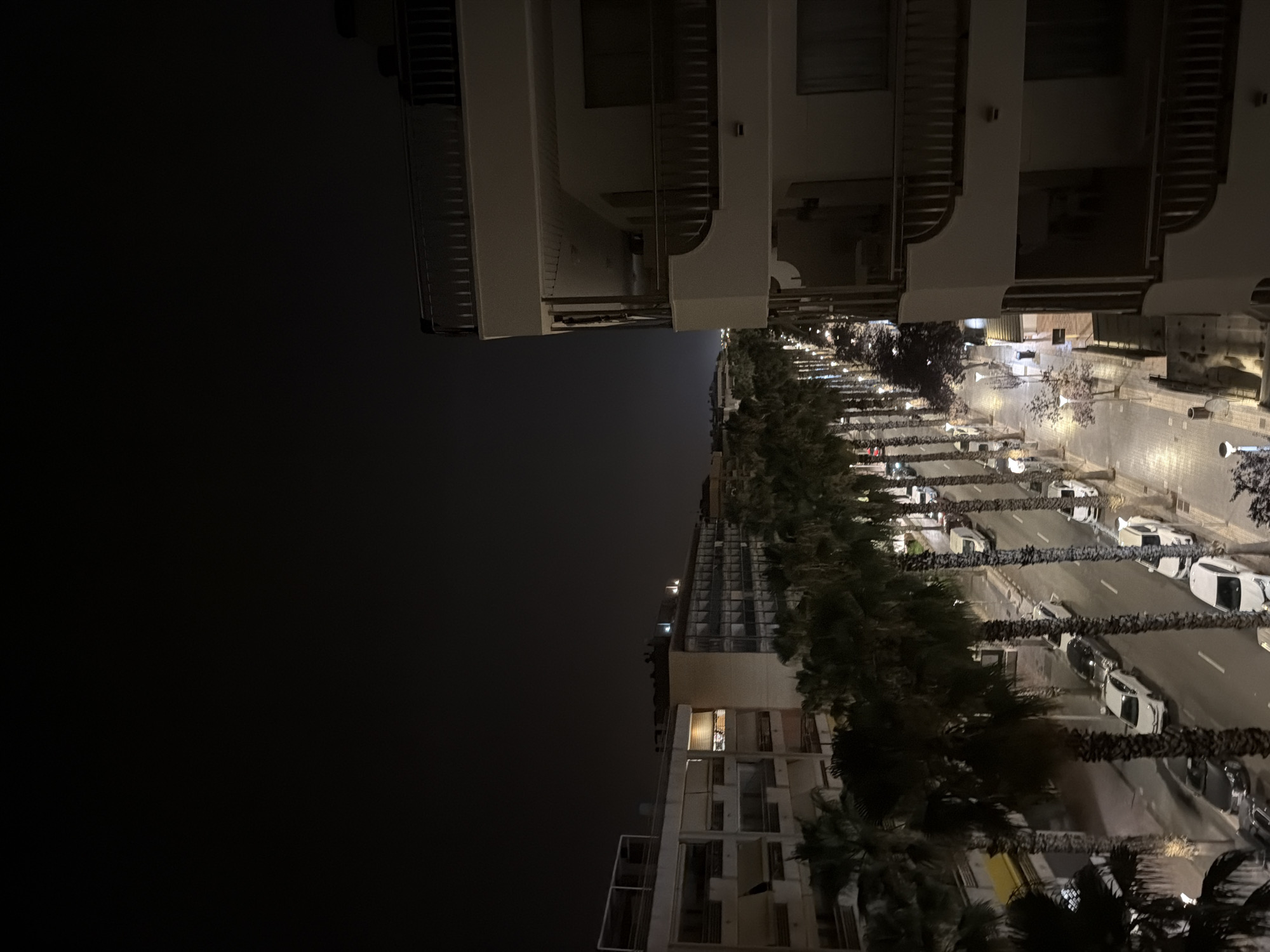 The image depicts a nighttime scene on a street lined with palm trees in Salou, Spain. The perspective is from an elevated position, looking down at the street and adjacent buildings. The street is illuminated by streetlights, casting a warm glow on the pavement. Several cars are parked along the sides of the street, and some are in motion.
On the left side of the street, a row of palm trees creates a distinct visual boundary. Behind the palm trees, multi-story buildings with balconies and illuminated windows are visible. One building on the right is a modern, multi-story structure with balconies and railings. The sky above is dark, indicating nighttime.
The overall impression is of a well-lit street in a coastal town at night, likely a tourist area given the palm trees and the style of buildings. The scene is quiet, with no people clearly visible in the immediate foreground. The environment appears to be a typical urban streetscape.