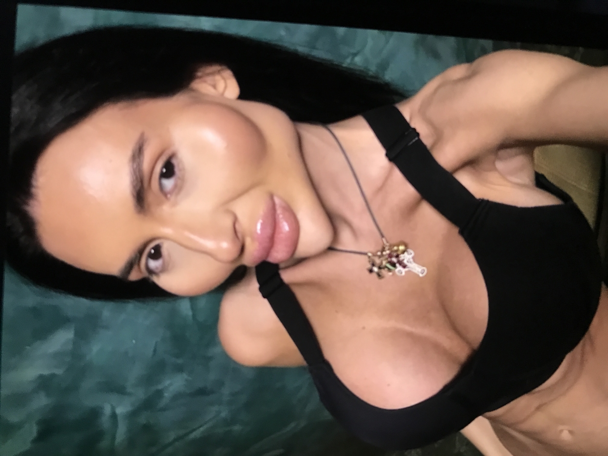 A woman with dark hair is angled towards the camera, wearing a black sports bra and a necklace with a colorful pendant. The background appears to be a textured green surface. It is difficult to determine the exact location in Donji Pobori, Montenegro, from this image.