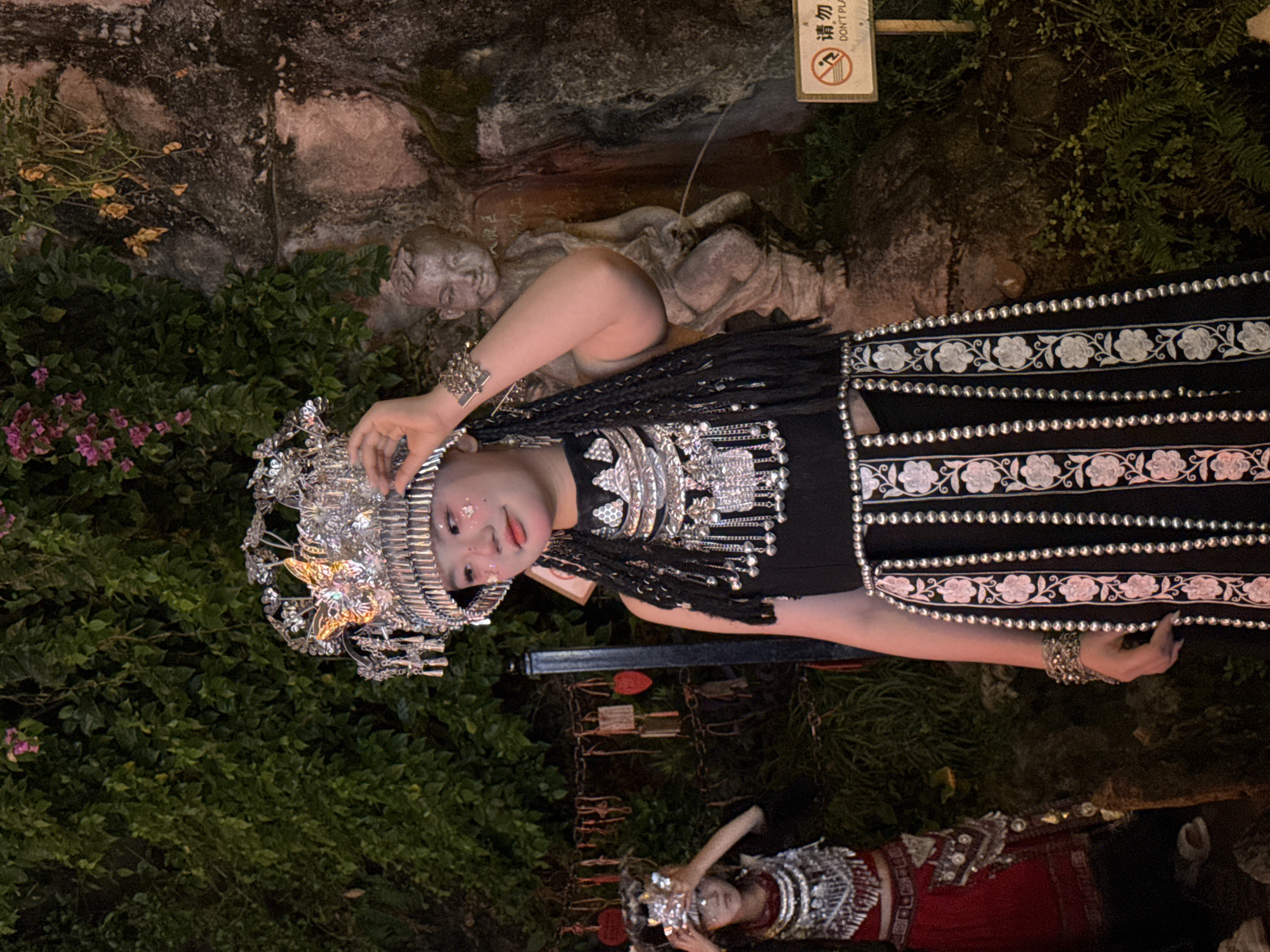 An outdoor nighttime scene in Yuzhong District, China, features two individuals adorned in traditional attire. The primary subject, a young woman, stands in the foreground wearing an elaborate silver headdress and a black dress embellished with intricate silver beadwork and floral embroidery. She has silver jewelry on her wrists and neck, and glitter on her cheeks. To her left, a child in a similar traditional costume, a red dress with silver embellishments, is partially visible. Behind the subjects, a stone statue of a young boy urinating stands against a rocky, moss-covered wall overgrown with green foliage and pink flowers. A chain with various inscribed tags hangs to the left of the woman. To the right, a small rectangular sign with red text and a prohibition symbol is affixed to a wooden post, indicating "DON'T PLAY" in English. The overall atmosphere is dimly lit, with artificial lighting highlighting the figures and the surrounding greenery.