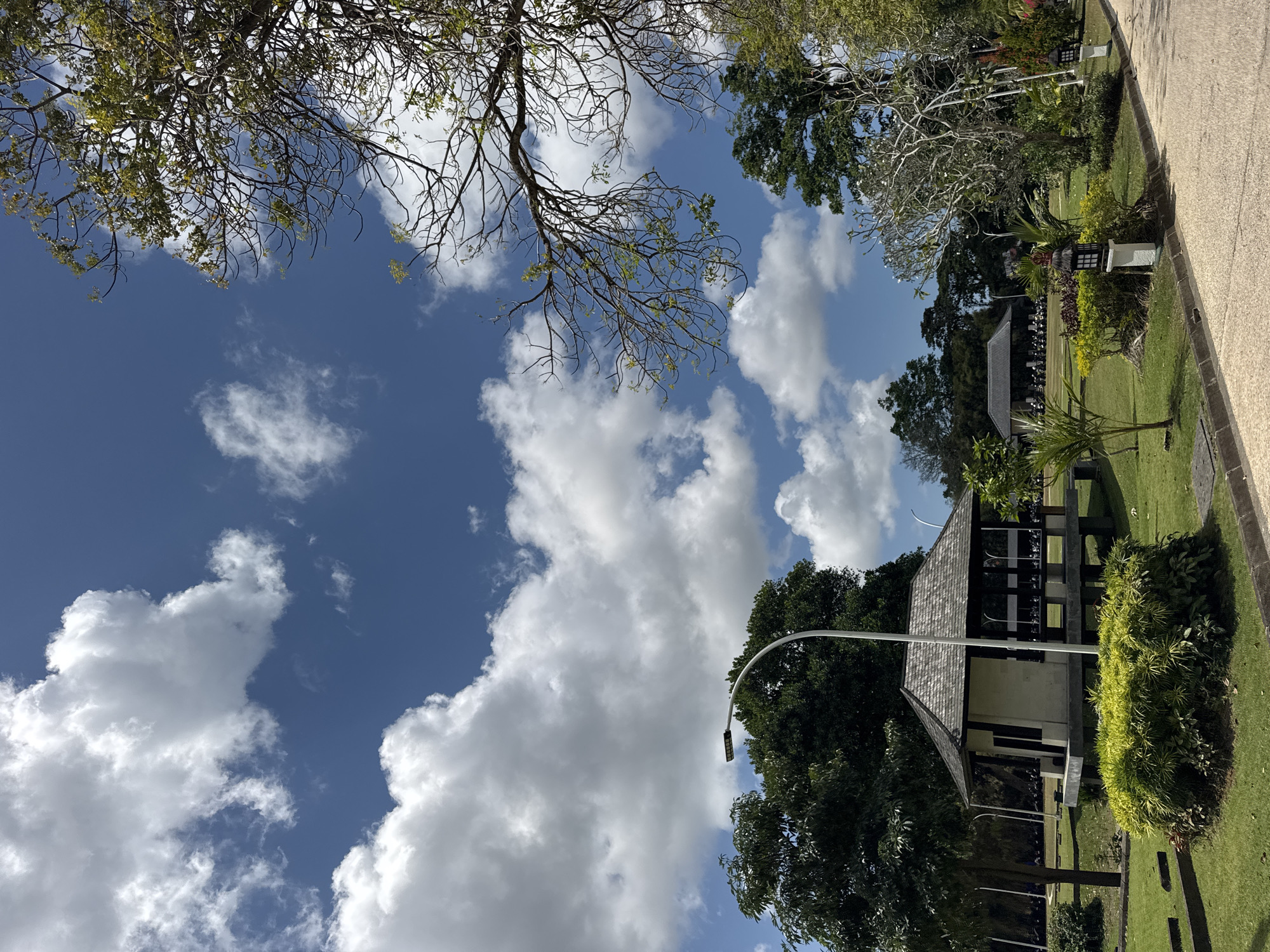 A medium shot captures a serene park scene under a vibrant blue sky with scattered white clouds. In the foreground, a paved walkway curves to the right, bordered by manicured green grass and low-lying shrubs. To the left, a small, traditional-style pavilion with a grey tiled roof stands elevated on stilts, surrounded by lush greenery. A modern street lamp with a rectangular LED fixture arches over the pavilion. Further into the background, more trees and structures are visible, including what appears to be another pavilion or building with a darker roof, and a row of parked motorcycles. The overall impression is one of a well-maintained public space, likely in a tropical or subtropical region. This scene is situated in Kuta Selatan, Indonesia, known for its beautiful beaches and cultural sites.