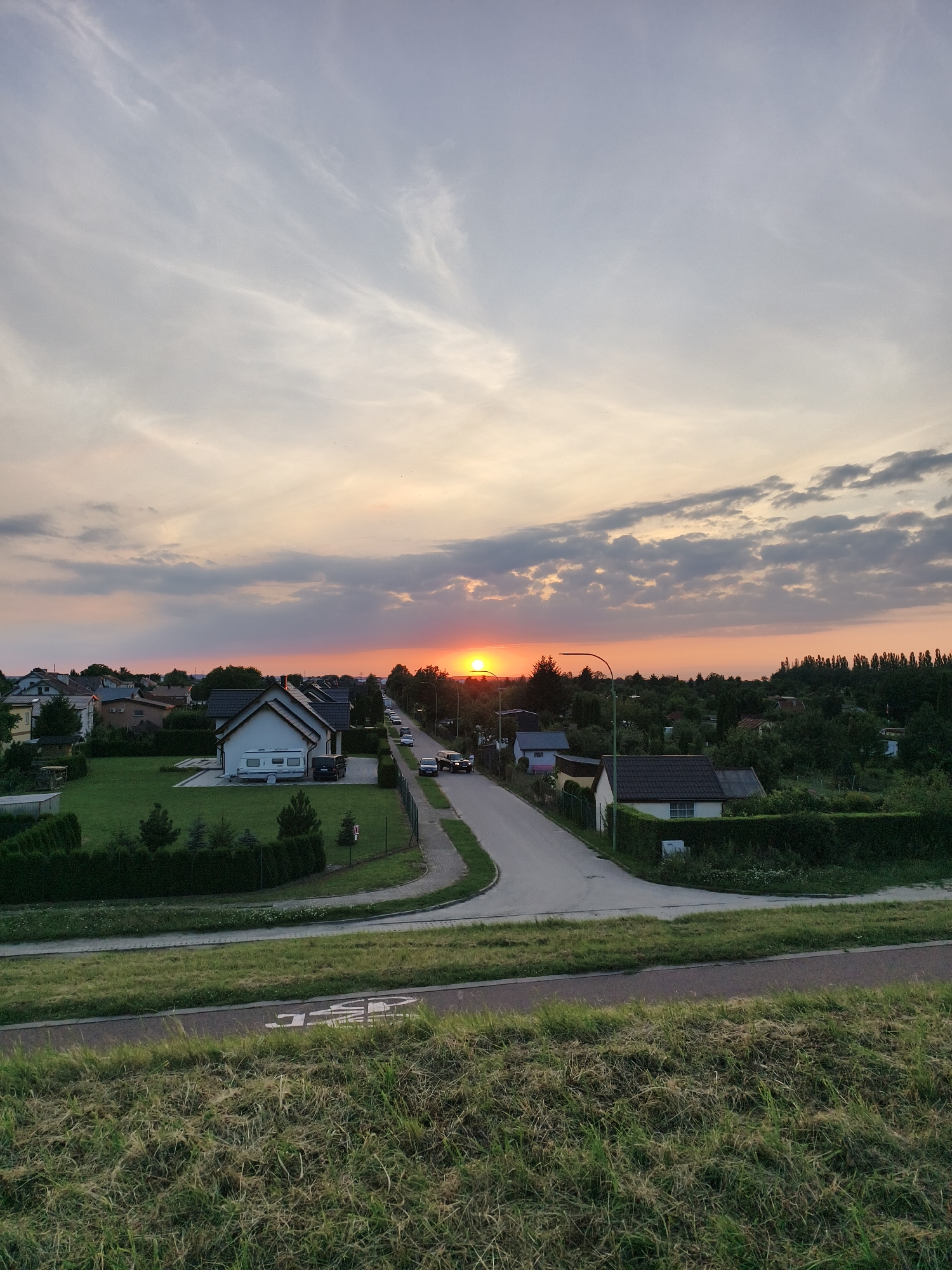 The image captures a suburban street in Tczew, Poland, during sunset. The sun, a bright orange orb, is visible low on the horizon, casting a warm glow across the sky and illuminating a scattering of clouds. A paved road winds through the scene, lined with modest residential houses and well-maintained green lawns. Several vehicles, including cars and a caravan, are parked along the roadside. Mature trees and hedges form natural boundaries between properties. In the foreground, a grassy embankment slopes down towards a bike path marked with white symbols. The overall atmosphere is serene and quiet, characteristic of an evening in a residential area.