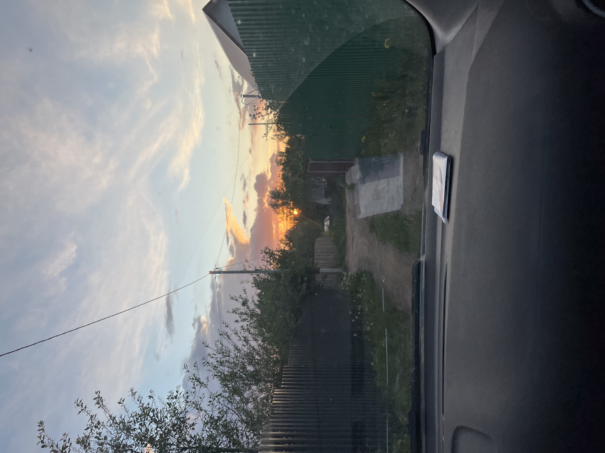 The image depicts a view from inside a vehicle, likely a car, through the windshield. Outside, a rural scene unfolds at sunset. A dirt road leads into the distance, flanked by green foliage, trees, and a corrugated metal fence on the left. In the background, the sun is setting, casting a warm glow through the trees. The sky is partly cloudy. A telephone pole with a wire is visible on the left side of the road. A cell phone is on the right side of the dashboard inside the vehicle. Based on the presence of buildings with the corrugated metal fences, the location is likely a rural part of a settlement in Chaikovsky, Russia. There is no people present in the image.