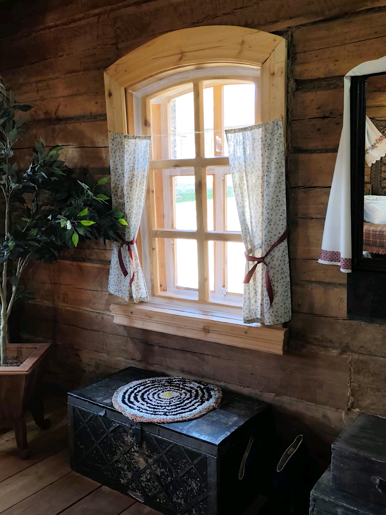 The image depicts an interior scene in a rustic, log-cabin-style room, likely within the Museum of Wooden Architecture in Taltsy, Irkutsk Oblast, Russia. A wooden window with a curved top is centrally located, adorned with light floral patterned curtains tied back with dark red ribbons. The walls are constructed of horizontal logs, showing a natural, textured finish. In front of the window, a dark, metal-banded chest serves as a surface for a circular, woven rug with a black, white, and yellow pattern. To the left of the chest and window is a tall, artificial green plant in a wooden planter. To the right of the window, a partial view of a mirror with a dark frame reveals a glimpse of embroidered fabric, possibly from a bedspread or curtain, on what appears to be a bed in the background. Another dark chest is partially visible in the lower right corner of the frame. The floor is made of wooden planks. The lighting suggests an indoor environment with natural light entering from the window. There are no people visible in the image.